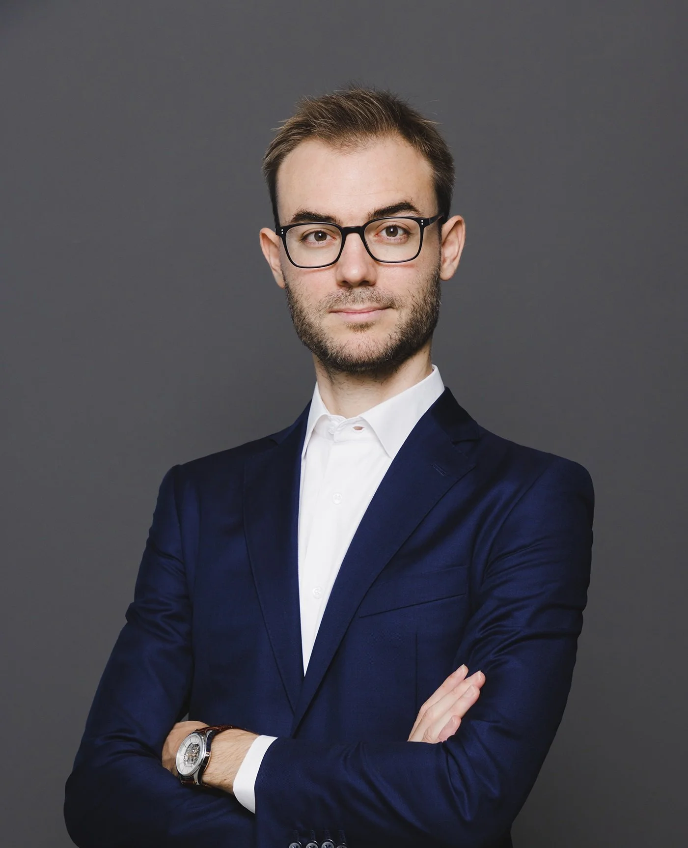 A young man with glasses, a beard, and neatly styled hair, wearing a dark blue suit and white shirt, crossing his arms against a gray background.