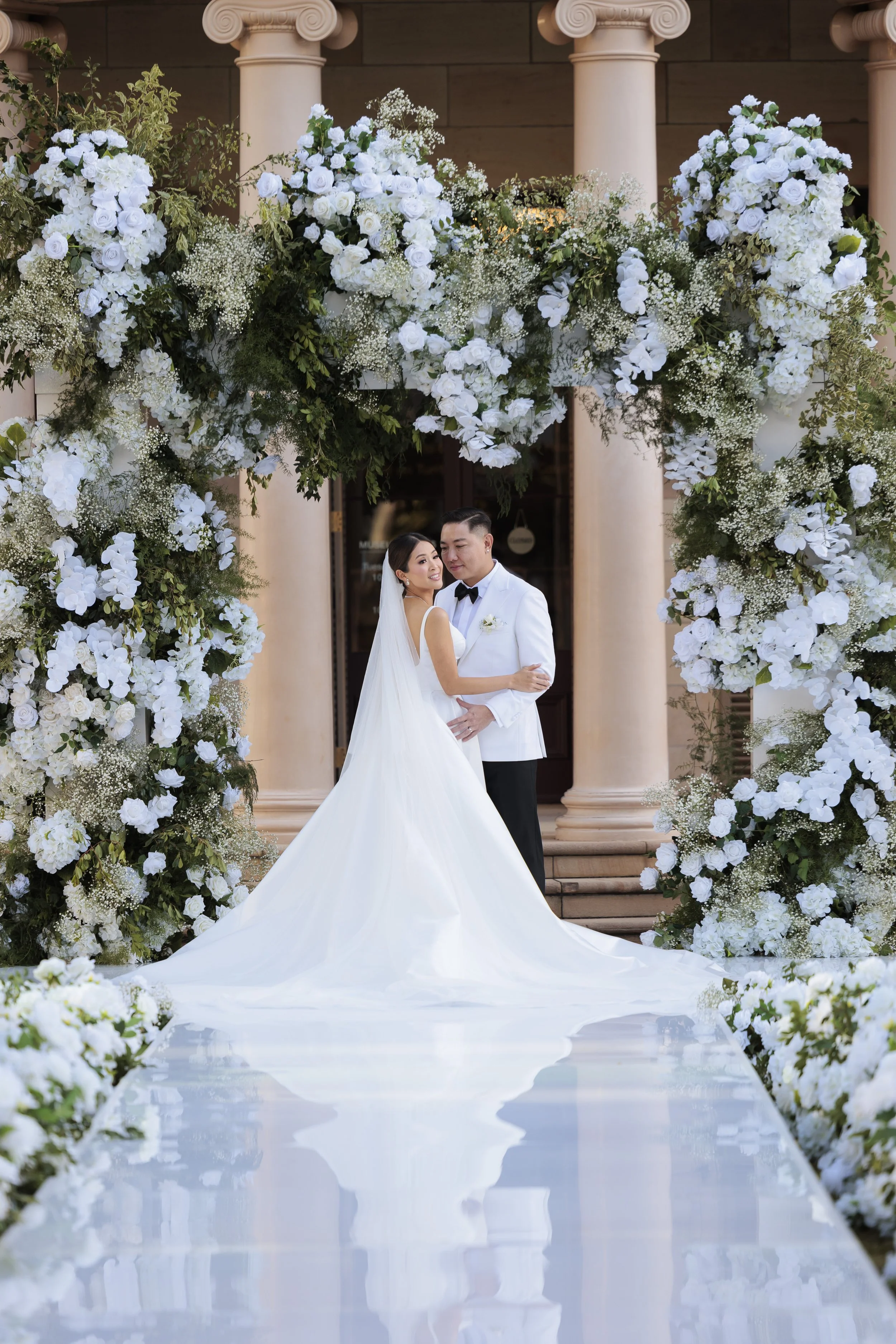 A bride and groom standing under a floral arch at their wedding, with cream-colored columns and a building in the background.