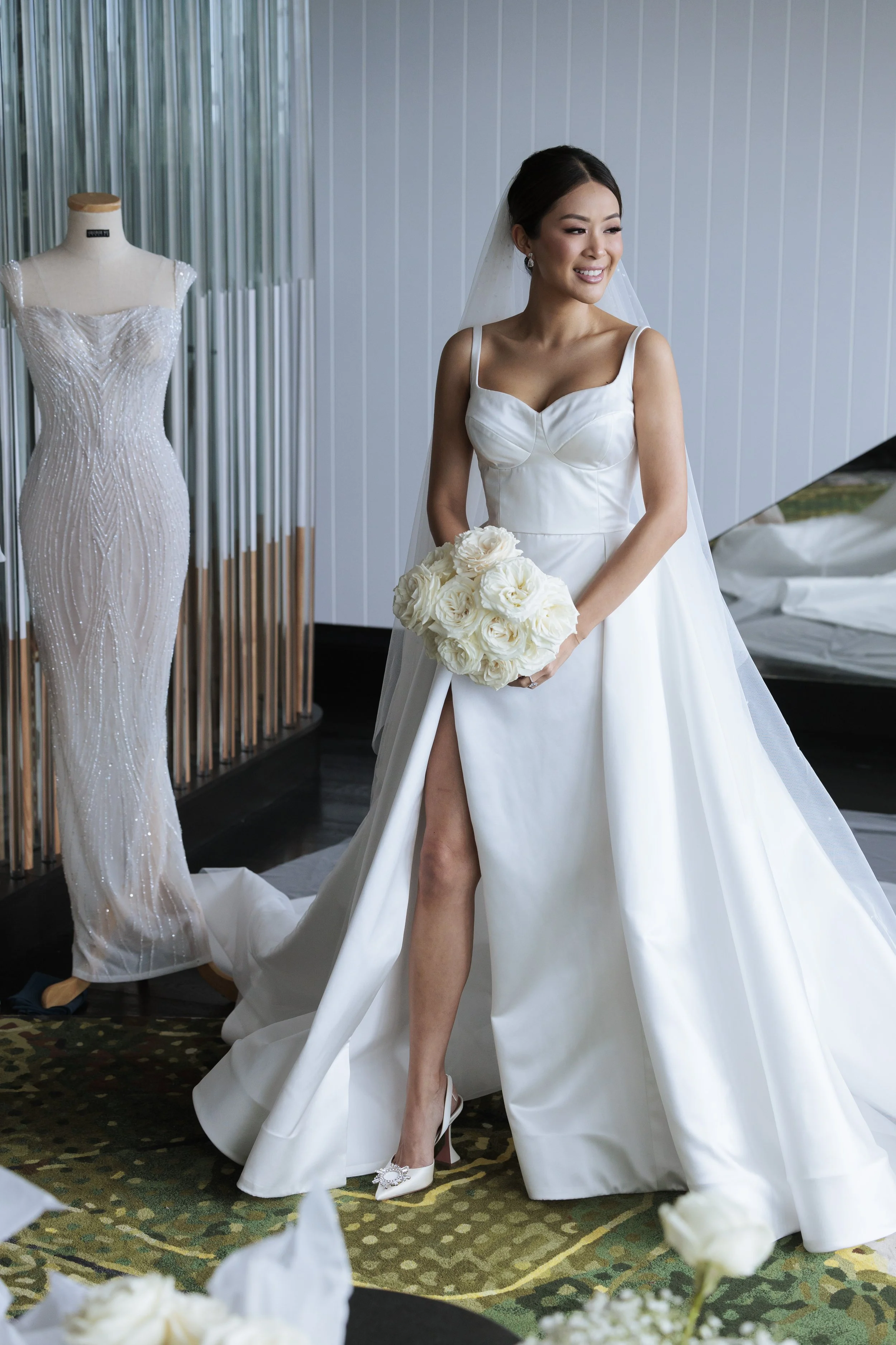 A bride in a white wedding gown holding a bouquet of white roses, smiling, standing in a bridal shop next to a mannequin wearing a beaded wedding dress.
