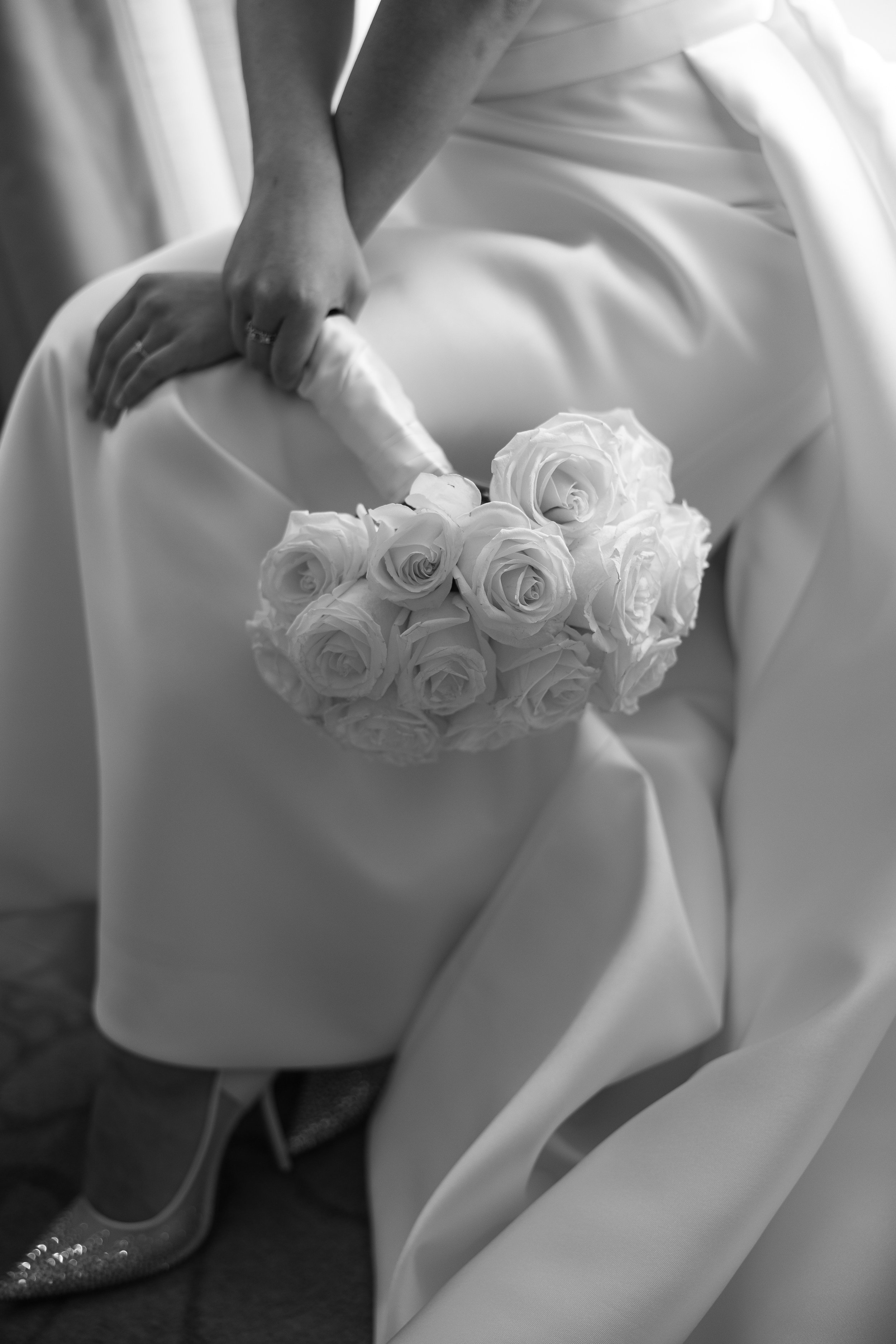 Close-up of a person's hand resting on their thigh, holding a bouquet of white roses, with a satin dress and sparkly shoe visible.