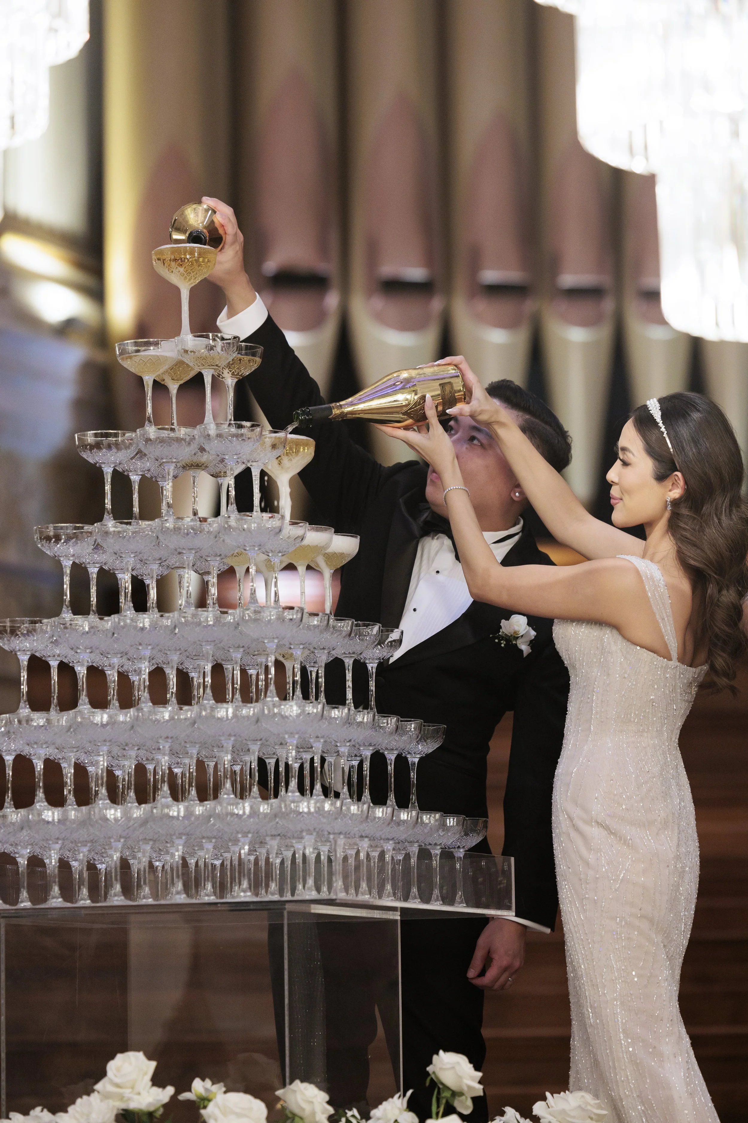 A newlywed couple building a champagne tower at their wedding reception, with the bride in a lace wedding dress and the groom in a tuxedo, pouring champagne to create the tower.