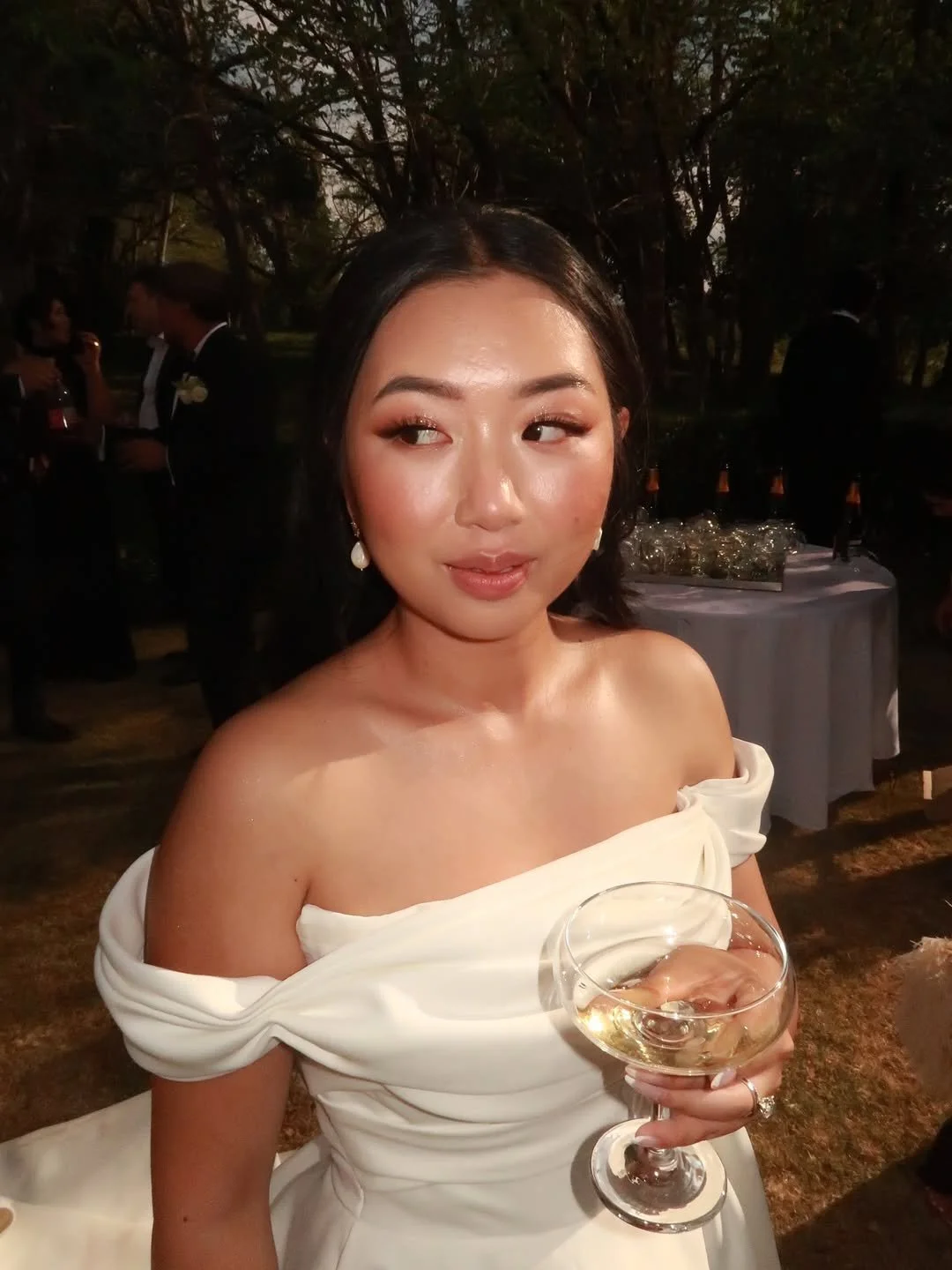 A woman in an off-shoulder white dress holding a glass of white wine at an outdoor event.