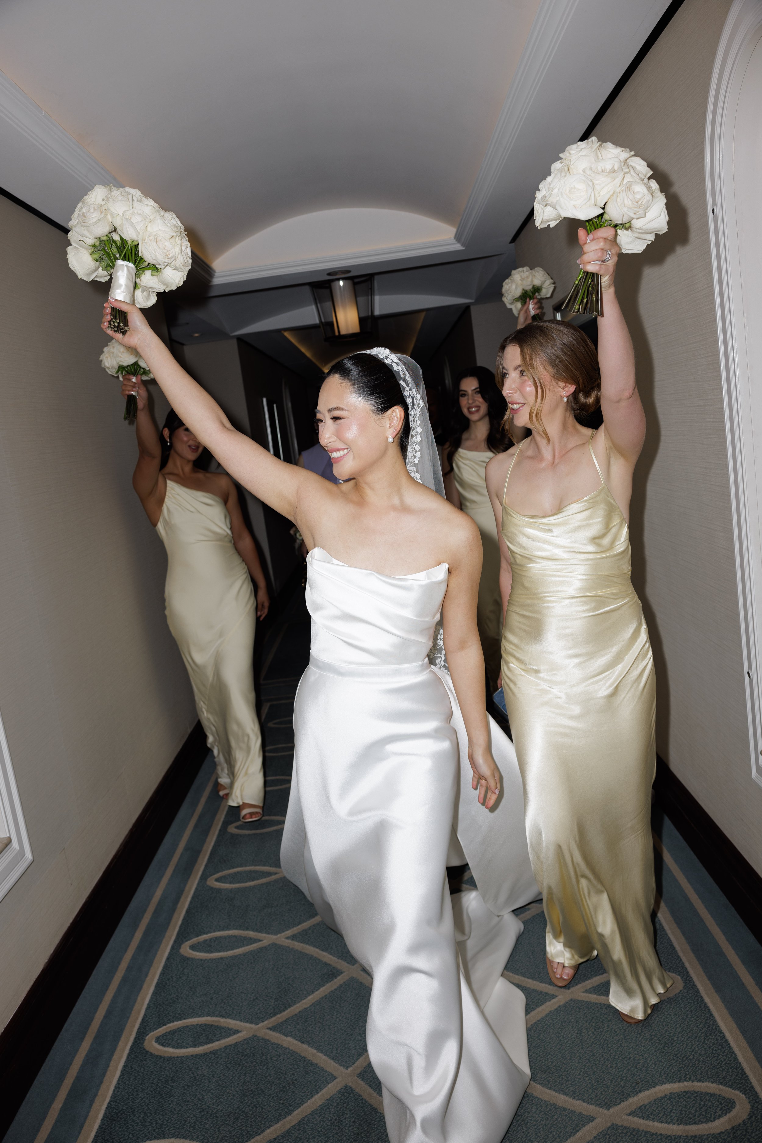 Women in wedding dresses and bridesmaids in satin dresses walking down a hallway holding bouquets of white roses during a wedding celebration.