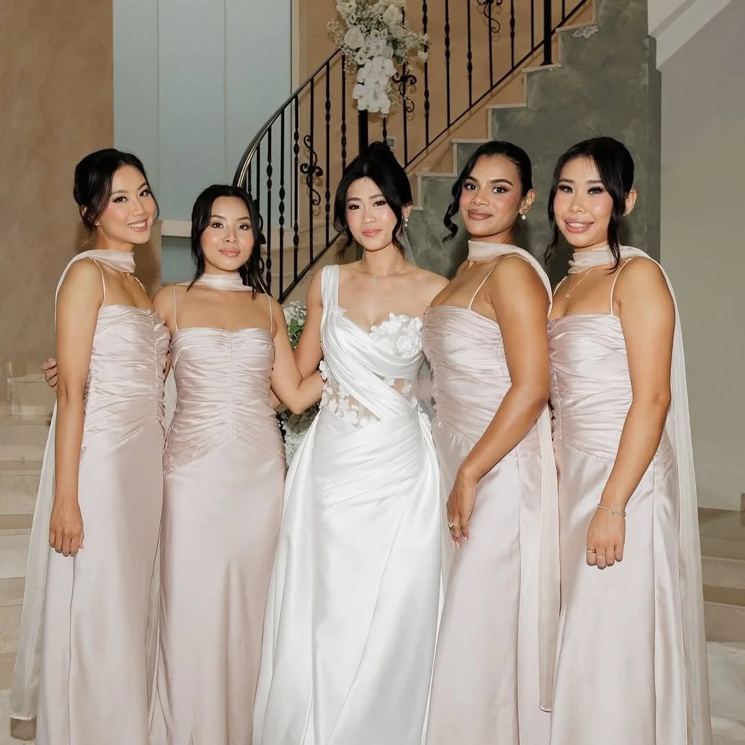 A bride in a white wedding gown standing with five bridesmaids in blush pink dresses inside a house near a staircase with floral decor.