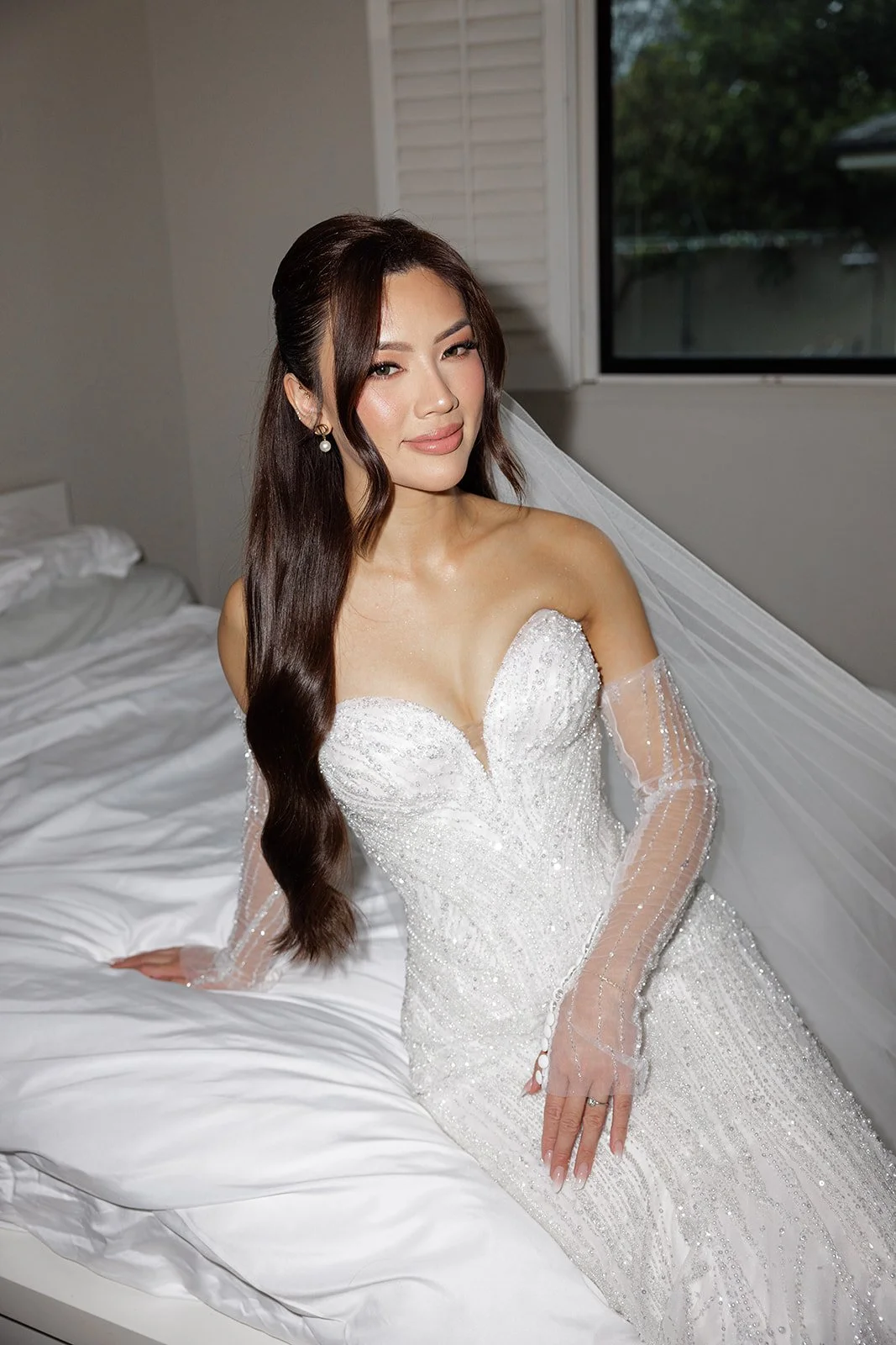 A woman in a white wedding gown sitting on a bed in a room with a window.
