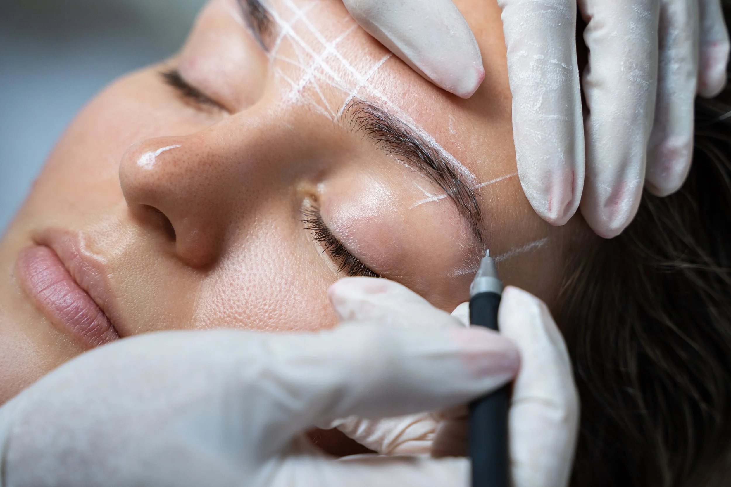 A person is lying down with their eyes closed while a close-up of their face shows they are getting a cosmetic tattoo procedure done on their eyebrow, with a tattoo machine in use and markings drawn on the skin.
