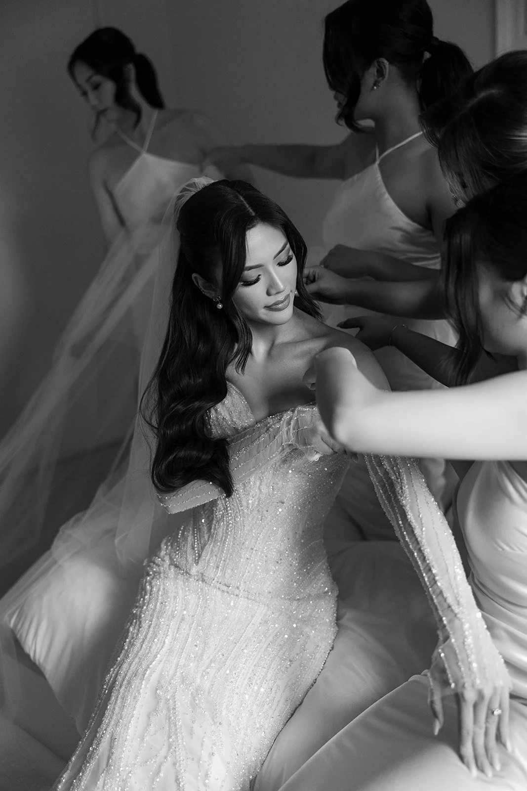 A bride getting ready with five women helping her, all wearing elegant dresses, in a room with soft lighting.
