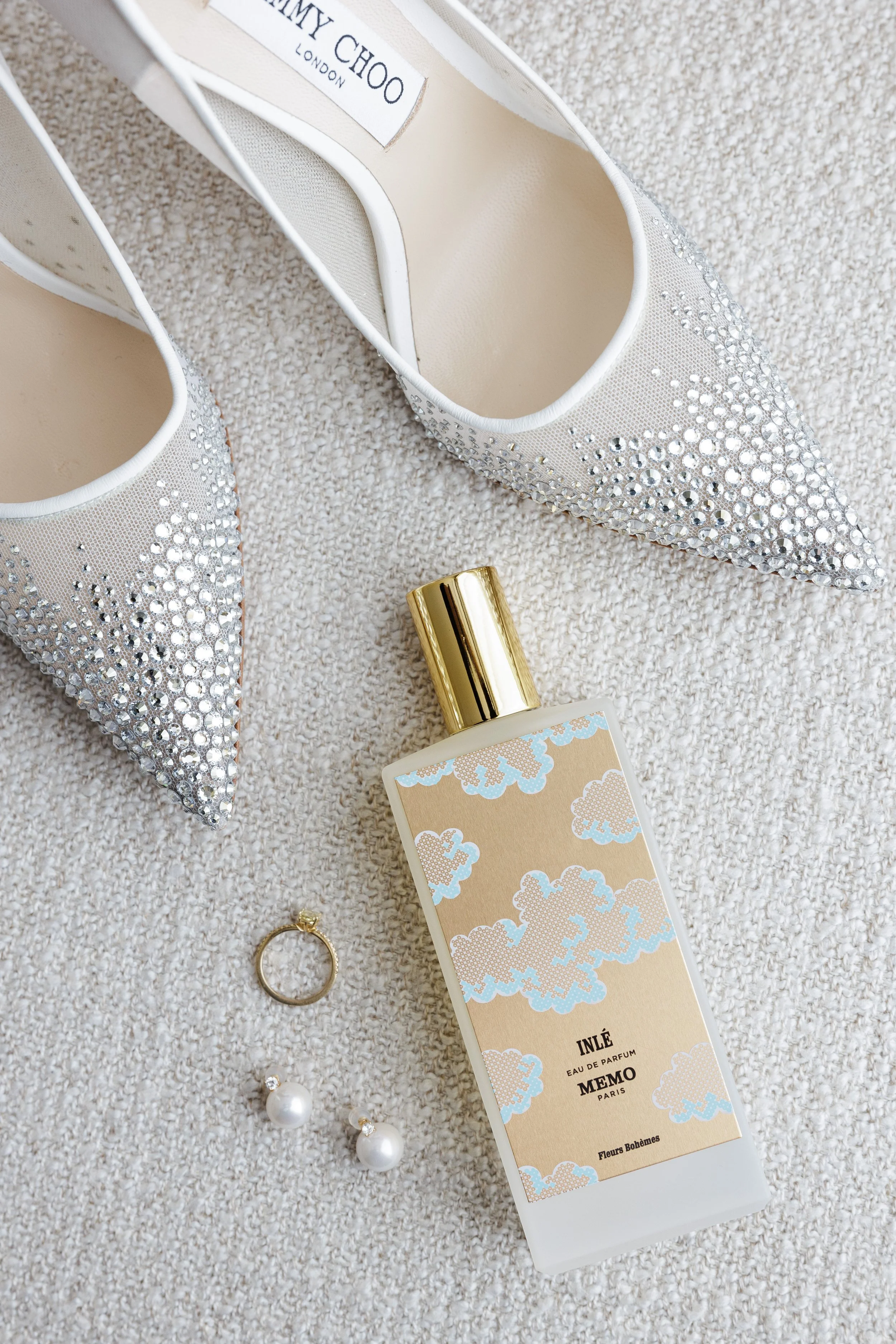 A pair of sparkling, pointed-toe high heels covered in rhinestones, a gold tube of perfume, pearl earrings, and a gold ring are arranged on a beige textured surface.