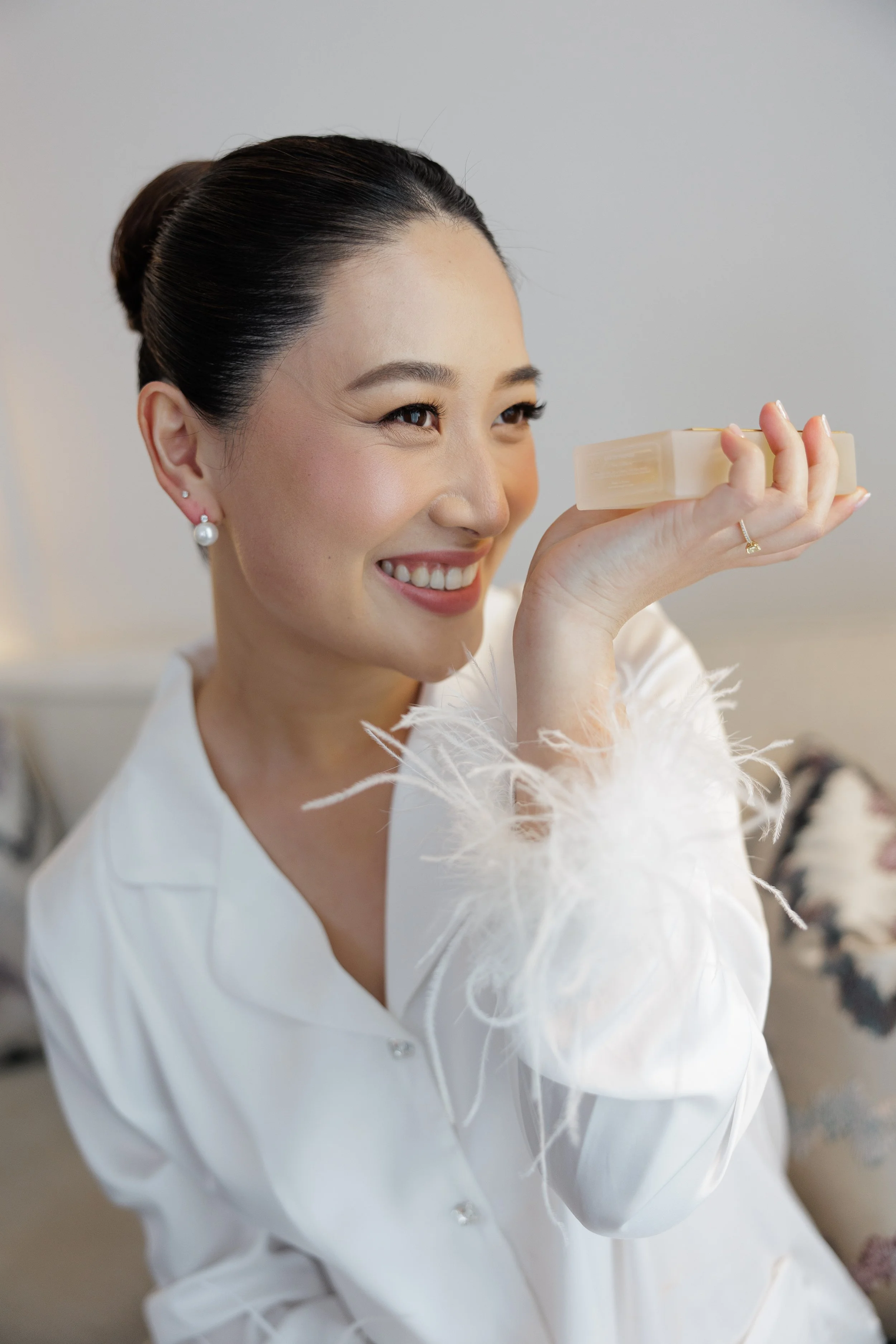 Smiling woman with dark hair in an elegant bun and pearl earrings holding a bar of soap or lotion close to her face, dressed in a white blouse with feather accents on the sleeves.