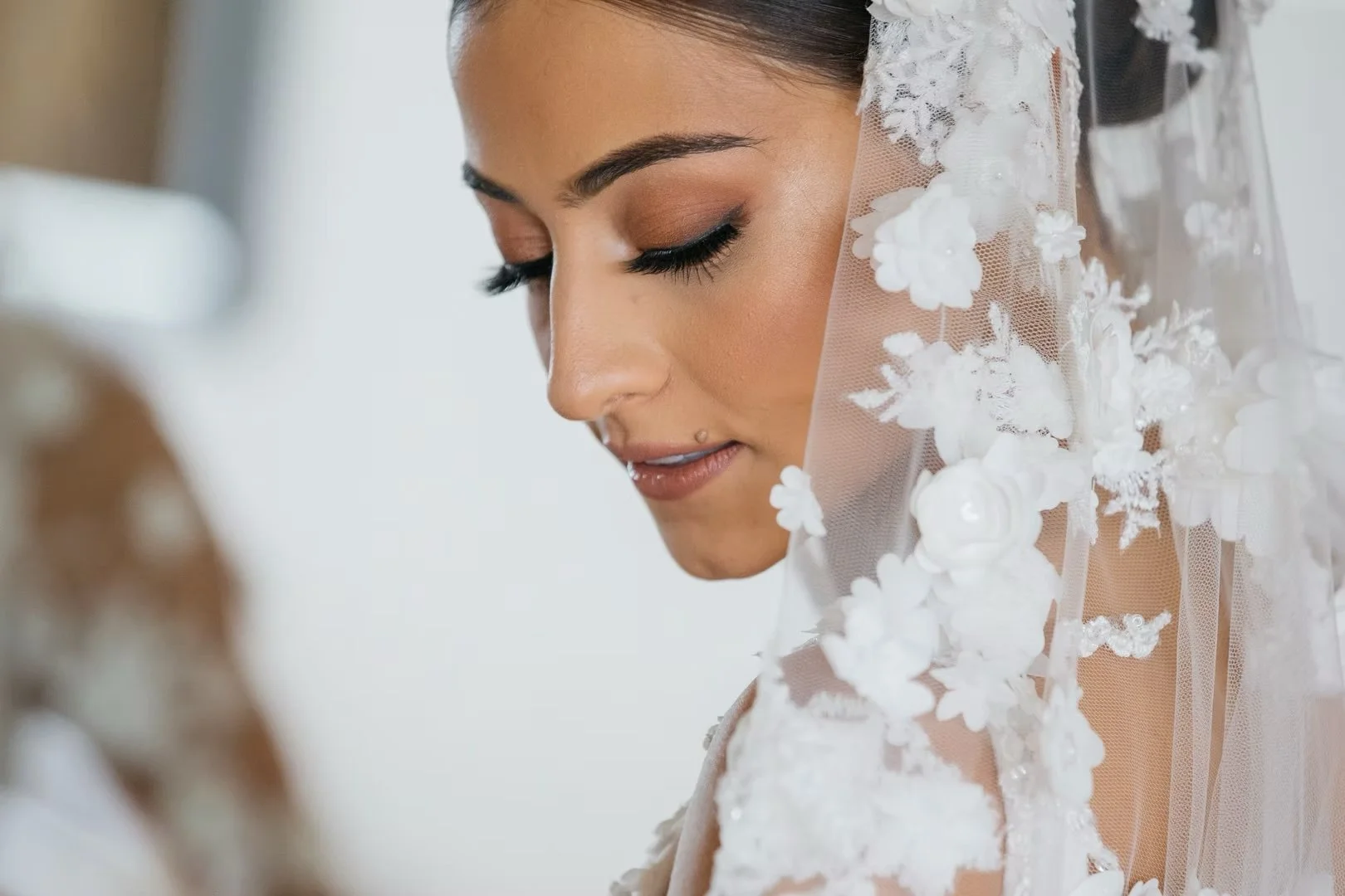 A bride with dark hair and makeup, wearing a white lace wedding dress and veil with floral embroidery, looking down with a peaceful expression.