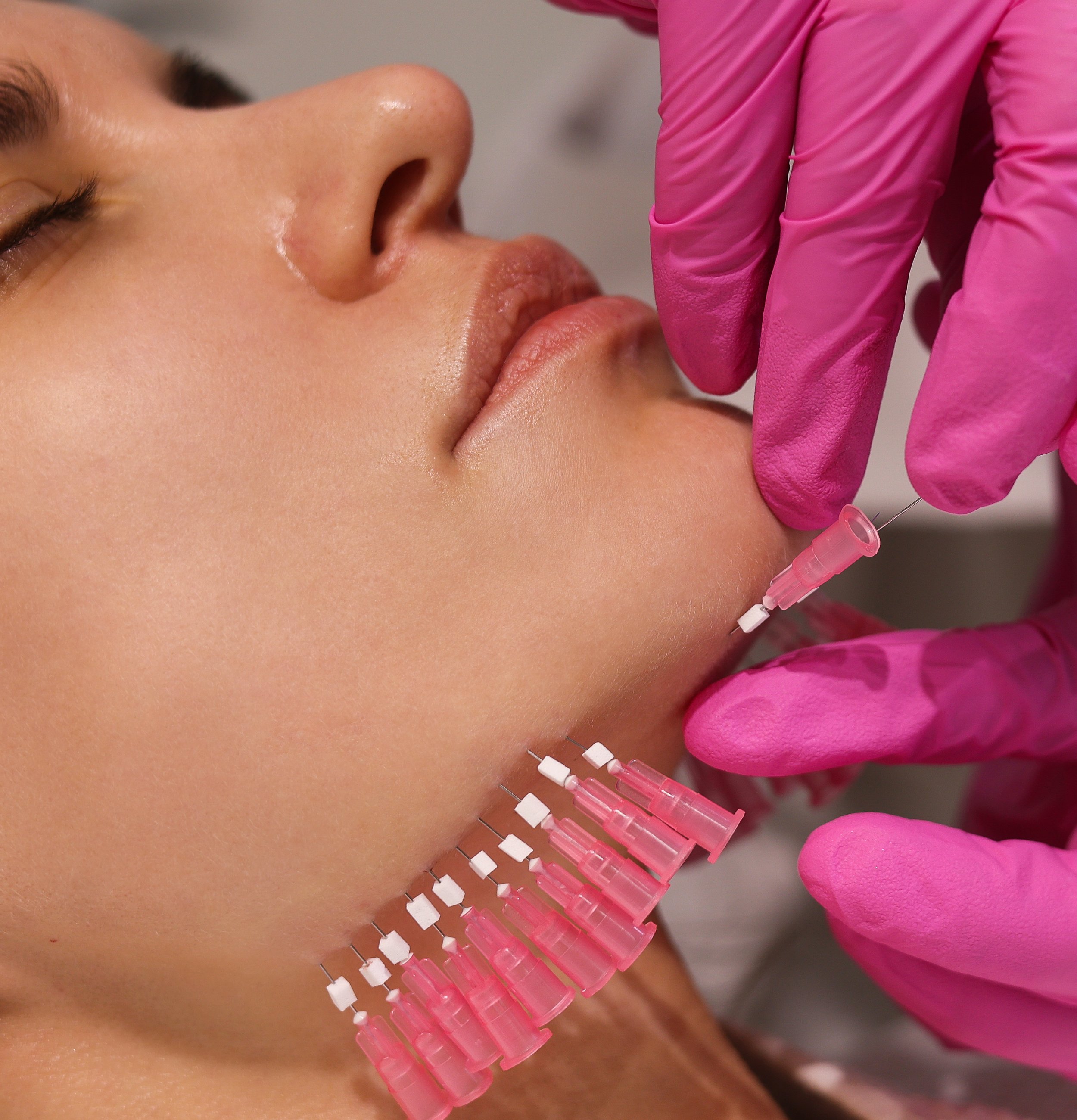 Close-up of a person's face with closed eyes receiving a vaccination shot in the upper arm from a healthcare worker wearing pink gloves, using a syringe with a needle.