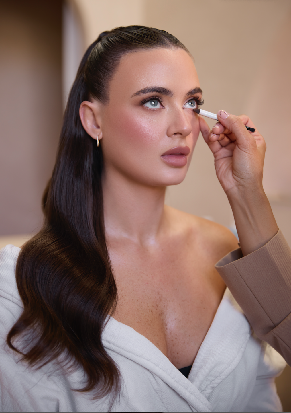 A woman with long, dark brown hair and blue eyes has makeup applied by a makeup artist who is using an eyeliner pencil to define her eyes.