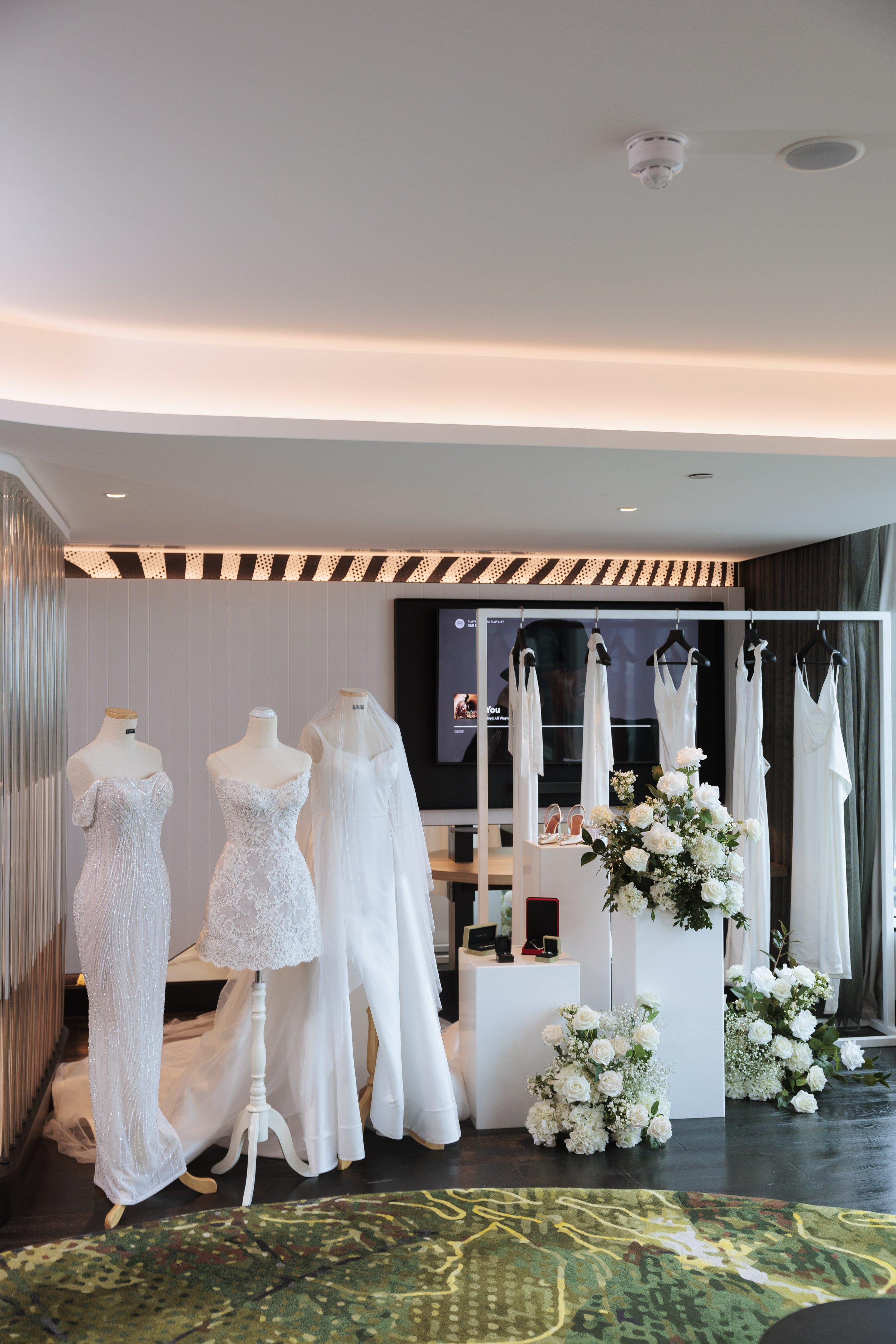 Display of wedding dresses and accessories in a boutique with floral arrangements and a patterned rug.