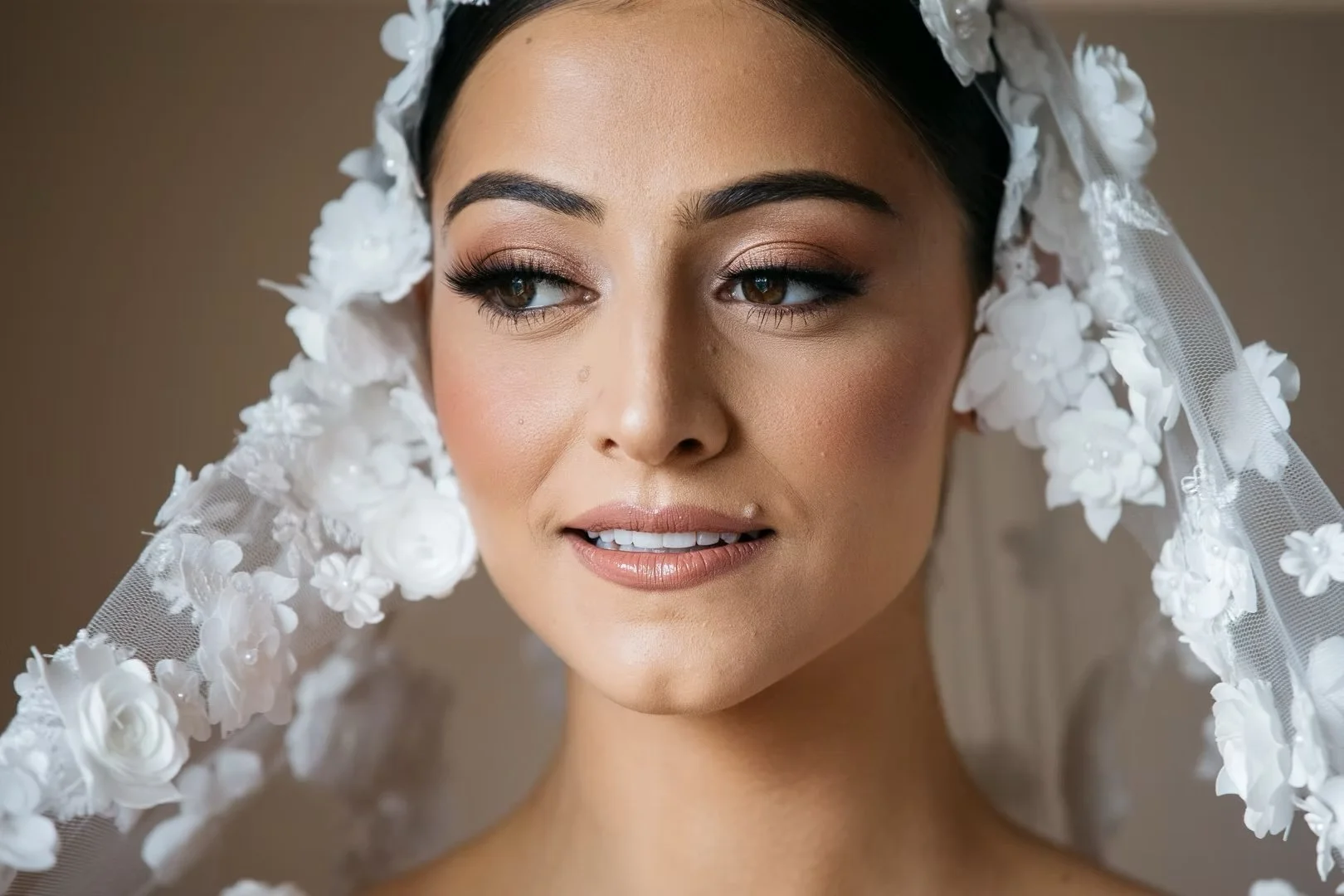 Close-up of a woman with makeup, wearing a wedding veil with white floral embellishments.