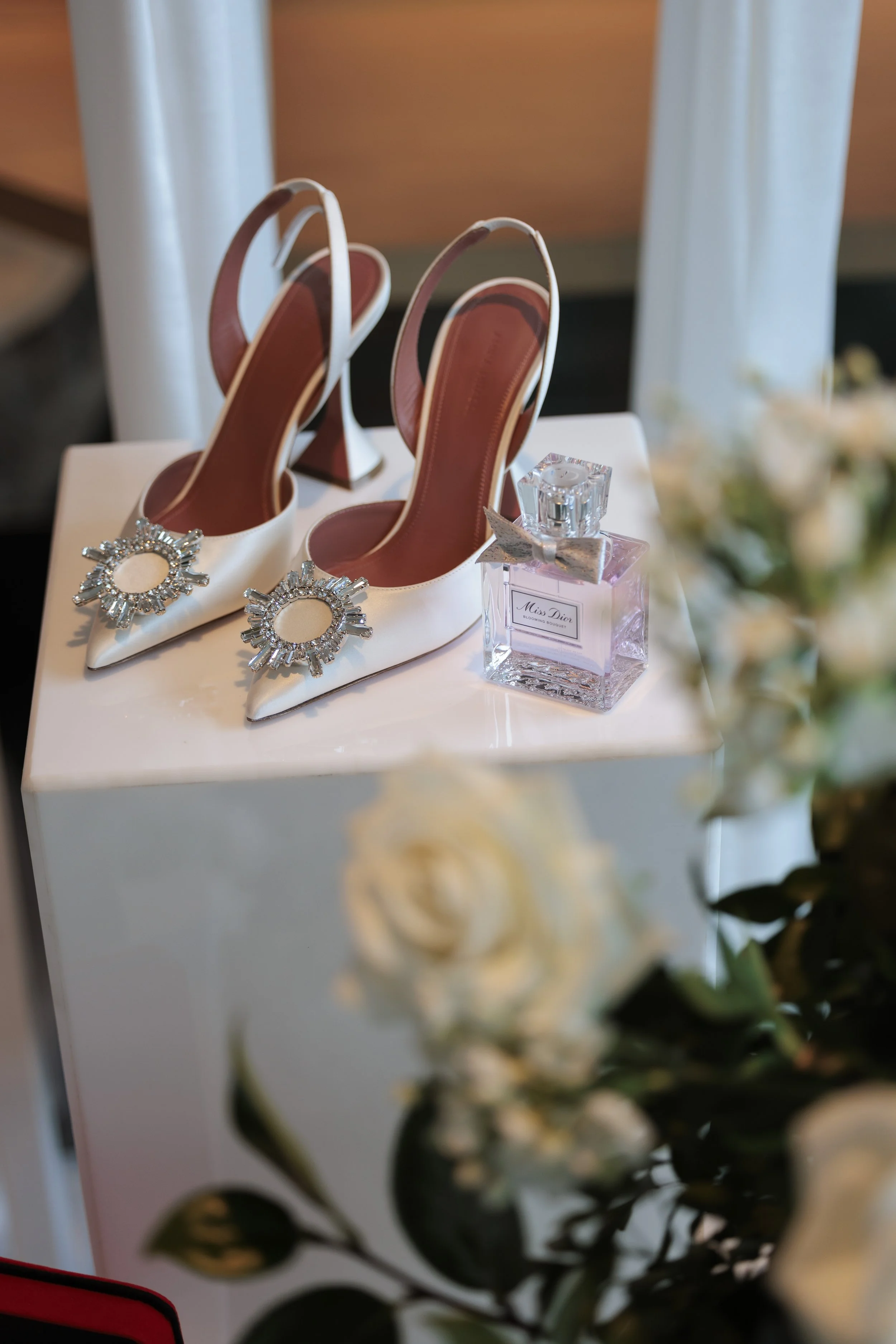 A pair of white high-heeled shoes with decorative gems on the toes, placed on a white surface alongside a bottle of Miss Dior Blooming Bouquet perfume.