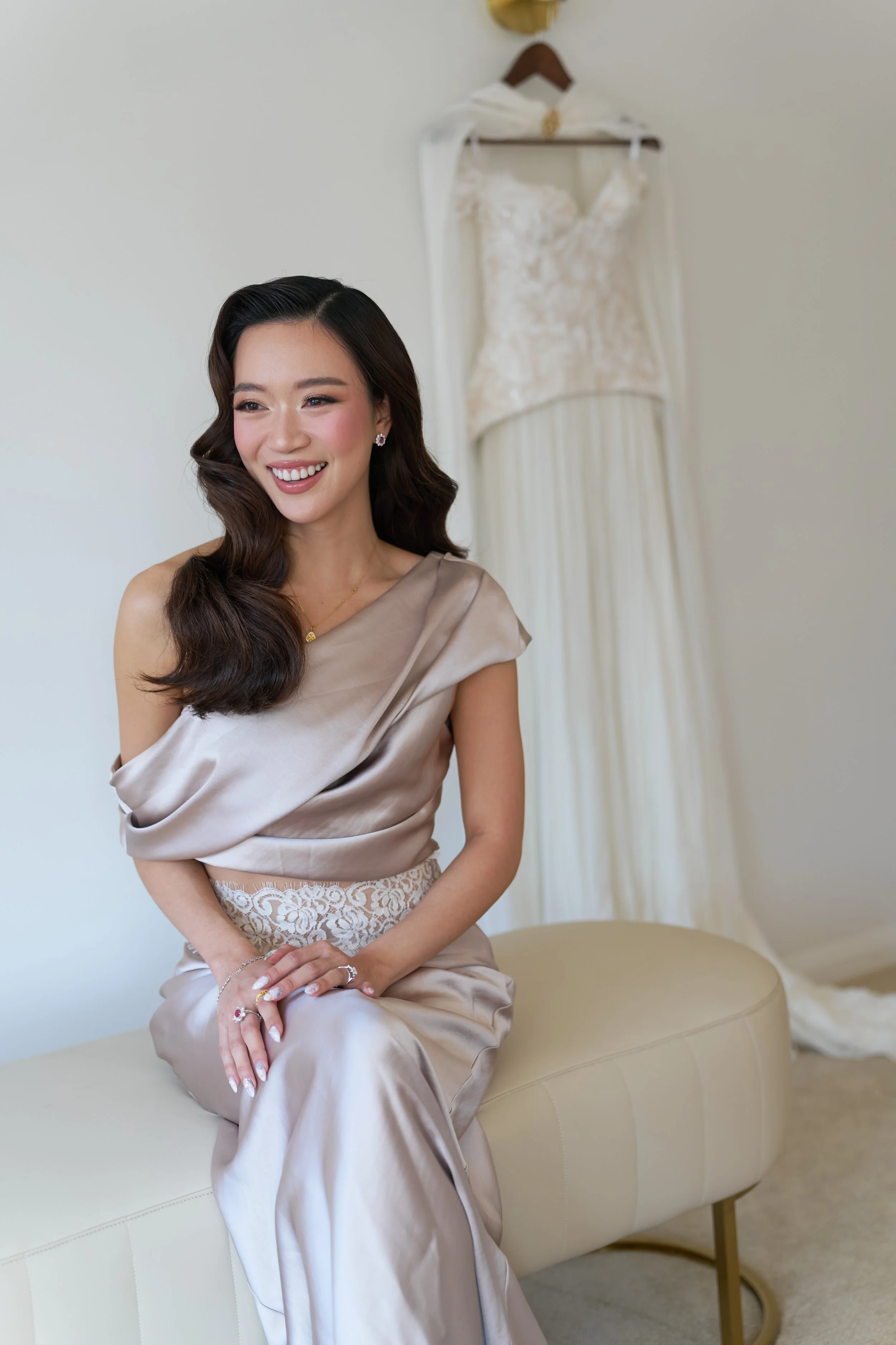 A smiling woman in elegant attire sitting on a cream-colored bench with a lace detail at her waist. Behind her on a wall, a wedding dress is hanging on a hanger.