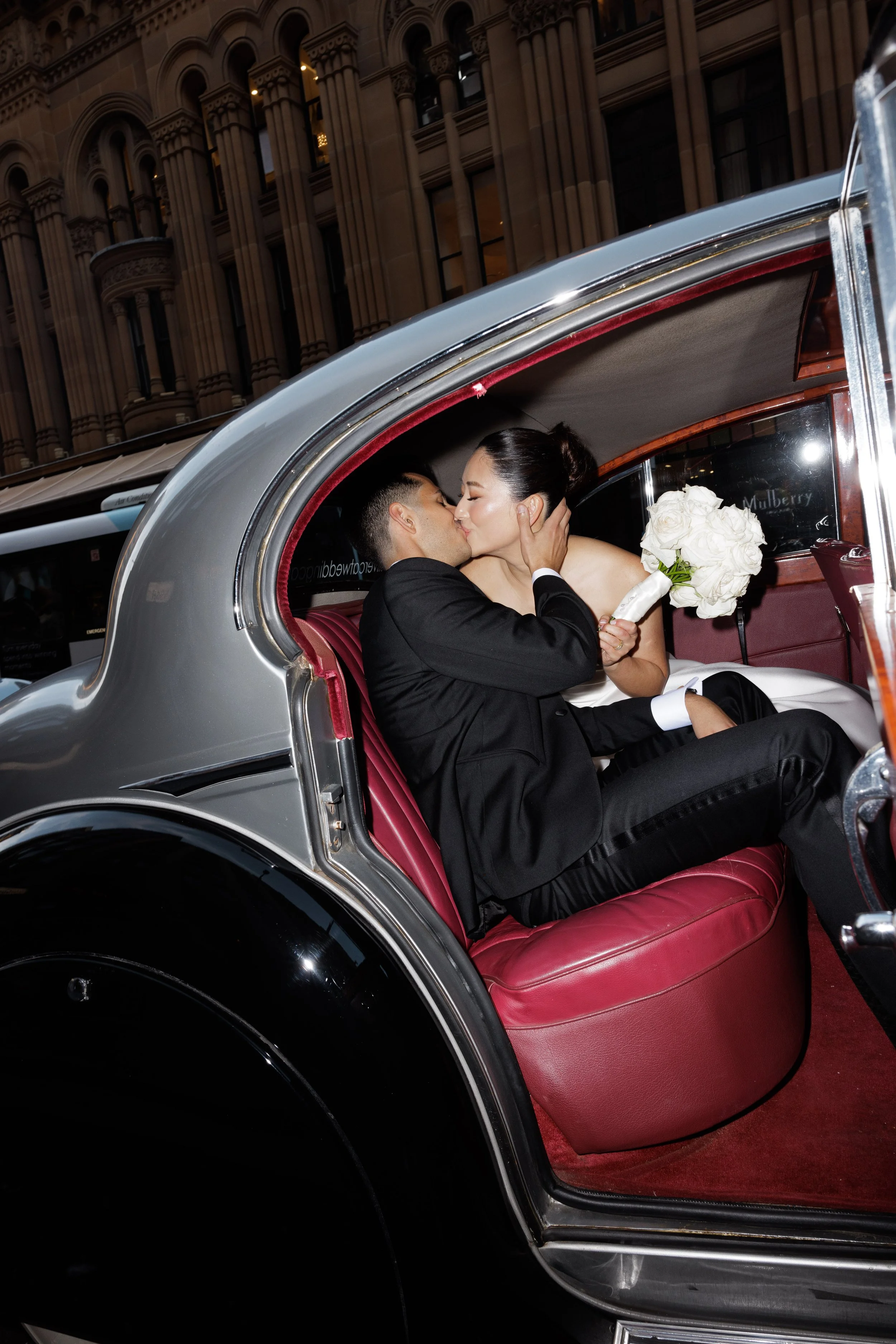 A man and woman sharing a kiss inside a vintage limousine, the woman holding a bouquet of white roses, with city buildings in the background.
