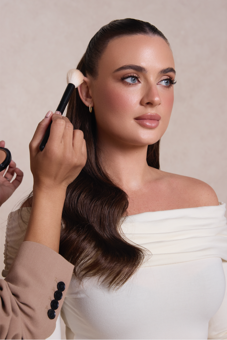 Woman with long brown hair and blue eyes is getting makeup applied, specifically blush or contour, with a large makeup brush by a makeup artist.