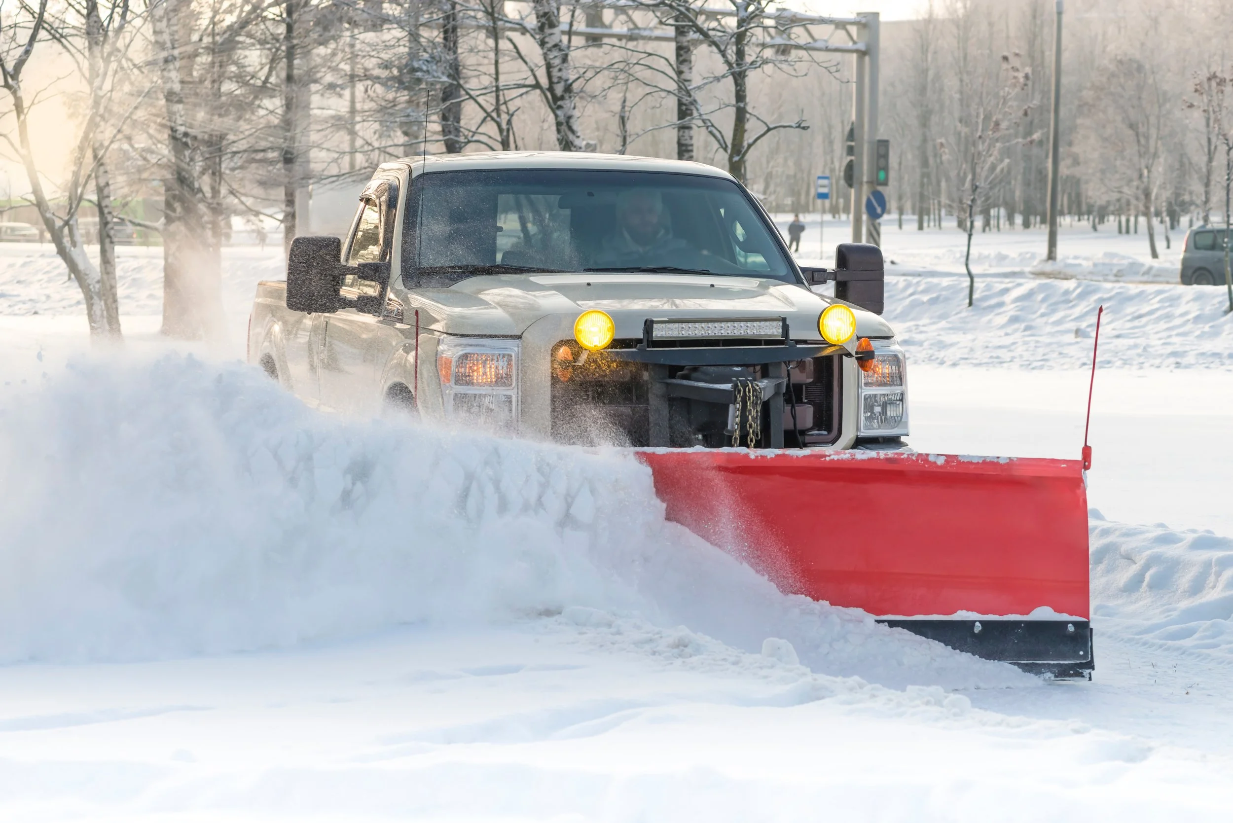 A snowplow clearing snow from a road in winter, with snow-covered trees and traffic lights in the background.