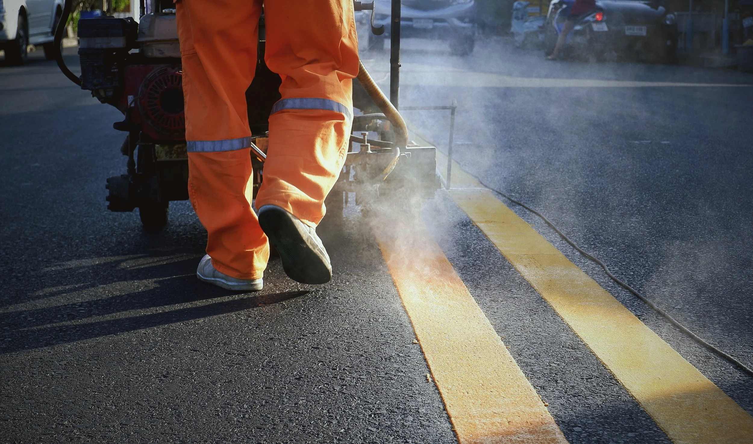 Worker in orange safety gear applying yellow road paint to create street markings on asphalt.