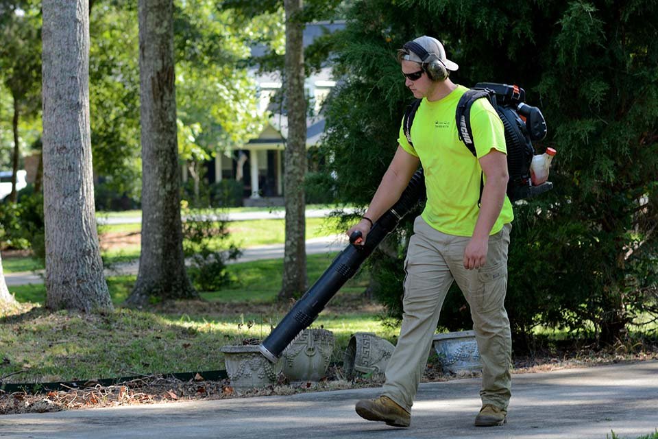 A person wearing a neon yellow shirt, beige pants, and a cap, using a leaf blower on a sidewalk in a residential neighborhood with trees and houses in the background.