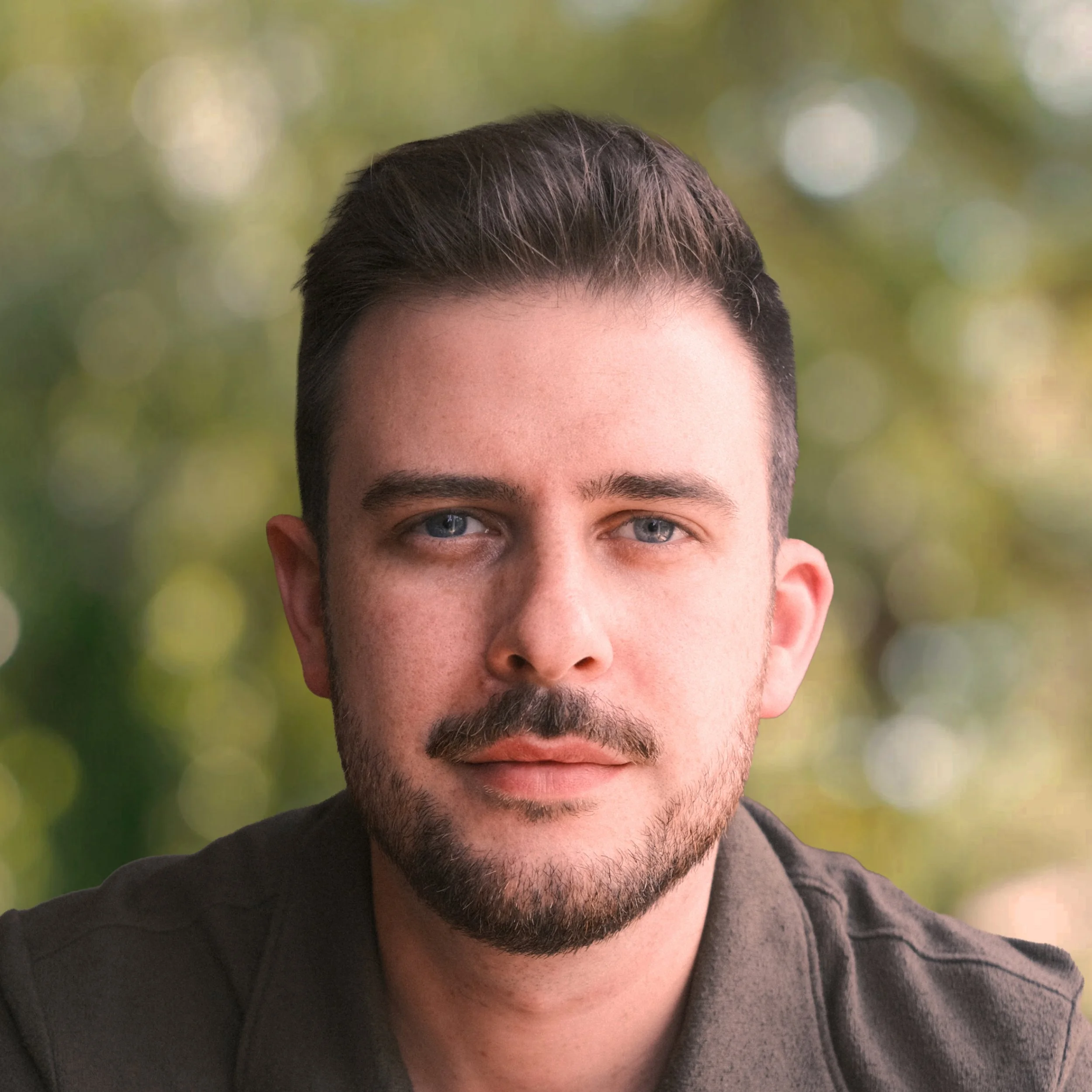 Close-up of a young man with short dark hair, light beard, and mustache, wearing a dark shirt, with a blurred outdoor background of trees and sunlight.