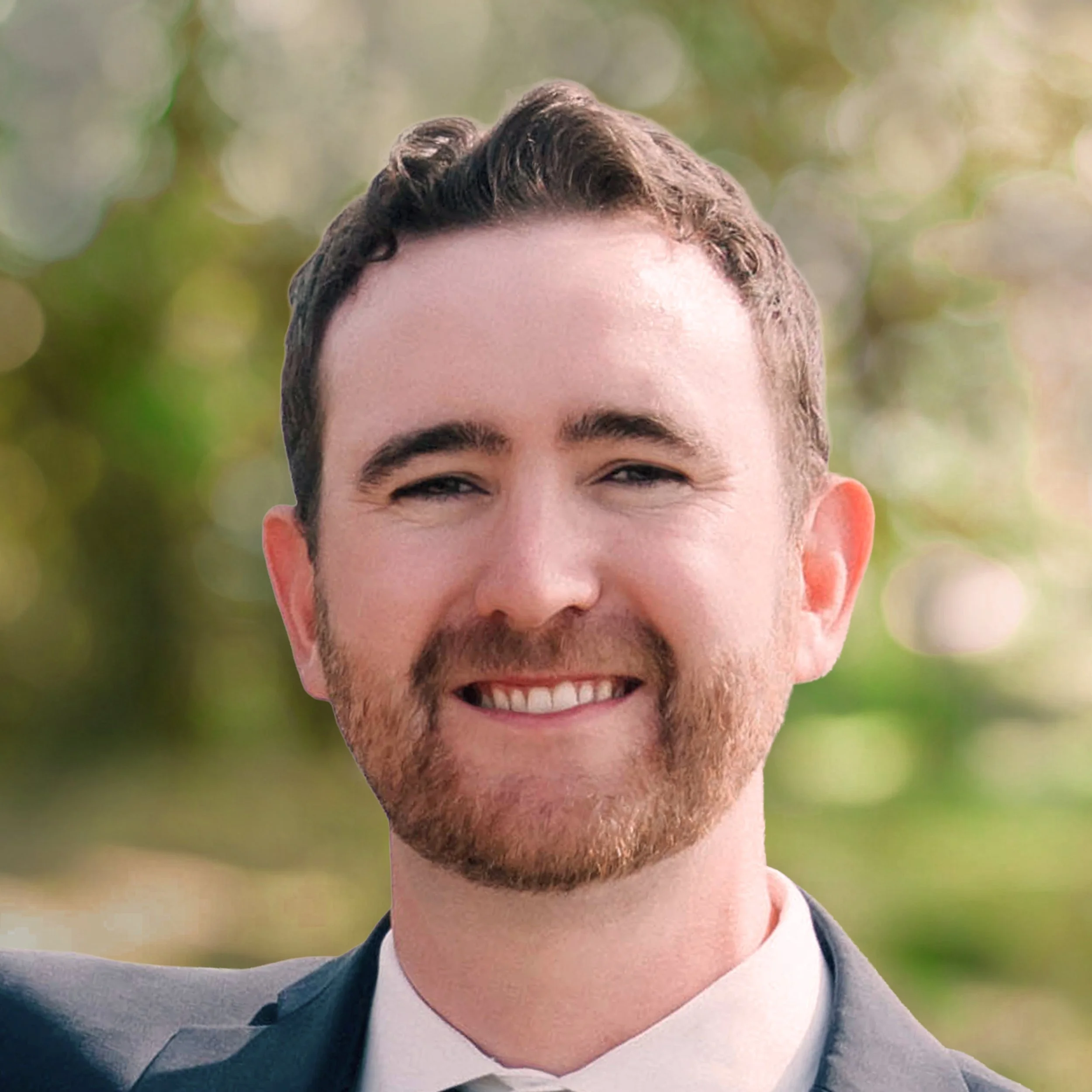 Close-up of a smiling man with short dark hair, beard, wearing a suit and tie, in front of a blurred green background.
