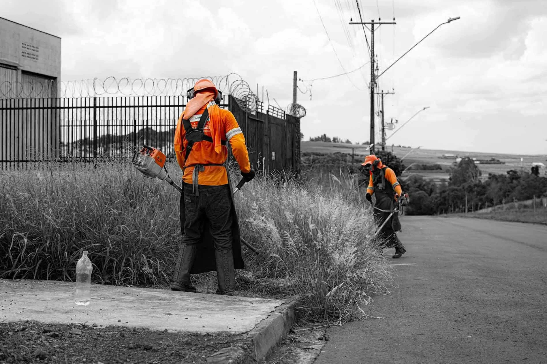 Two workers in orange uniforms and hats work along a roadside, one using a hedge trimmer and the other with a weed whacker, near a fence topped with barbed wire and old utility poles in a rural landscape.