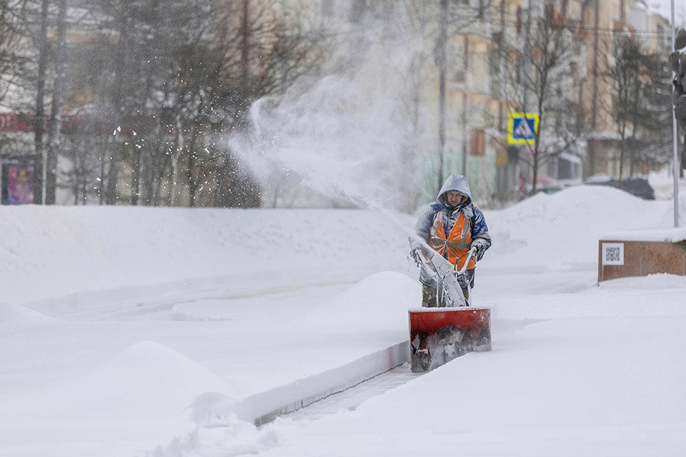 A person in a hooded jacket using a snowblower to clear snow from a sidewalk in a snowy city street.