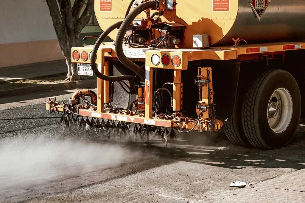 A street sweeper truck spray cleaning asphalt pavement on a city street with dust or water mist.