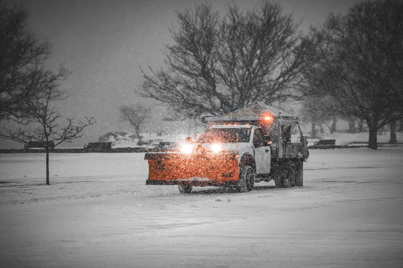A snowplow clearing snow on a road during a snowstorm in a park, with leafless trees in the background.