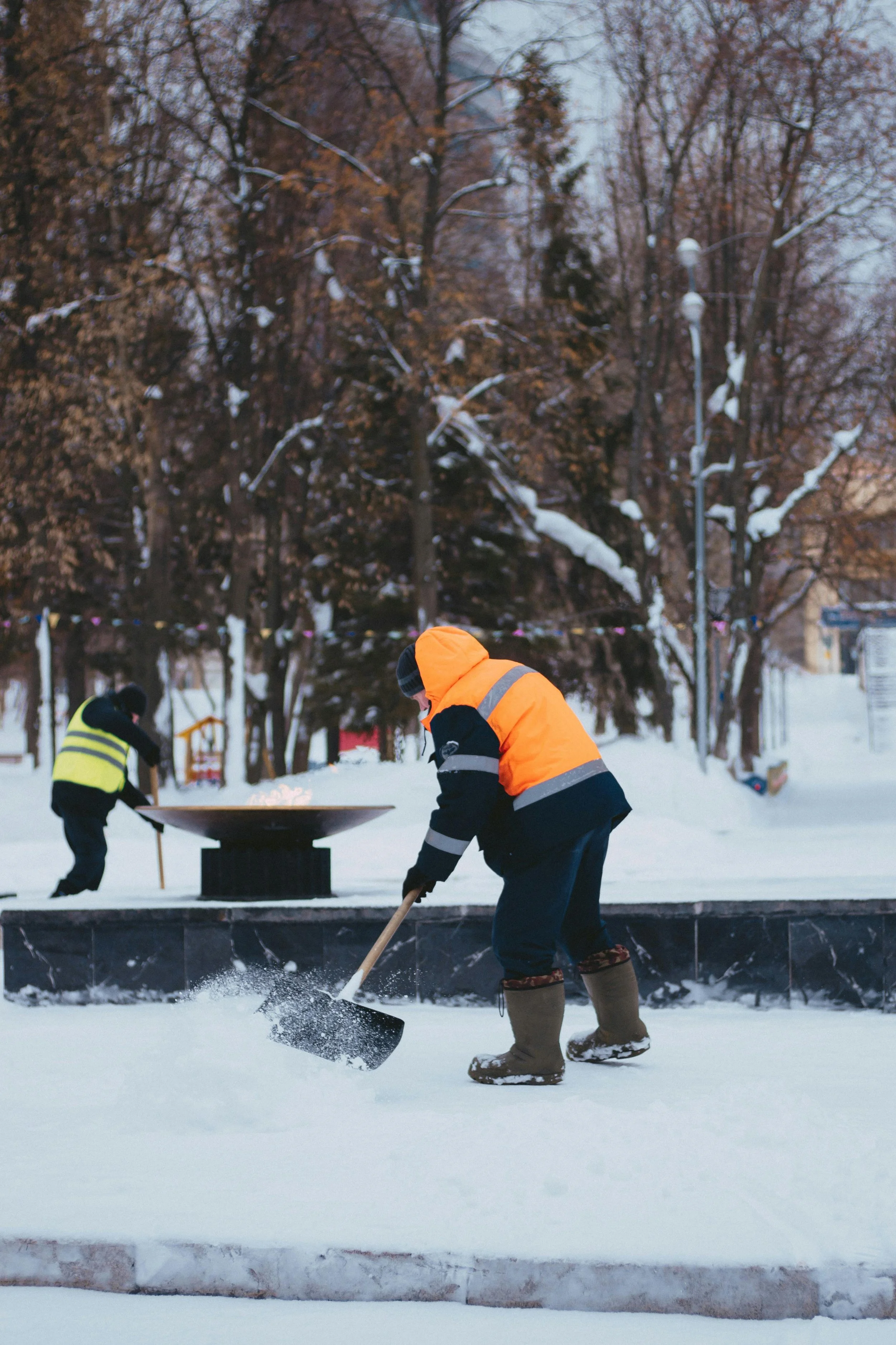 Two workers in winter clothing shoveling snow in a park during winter, with trees covered in snow and a burning fire pit in the background.