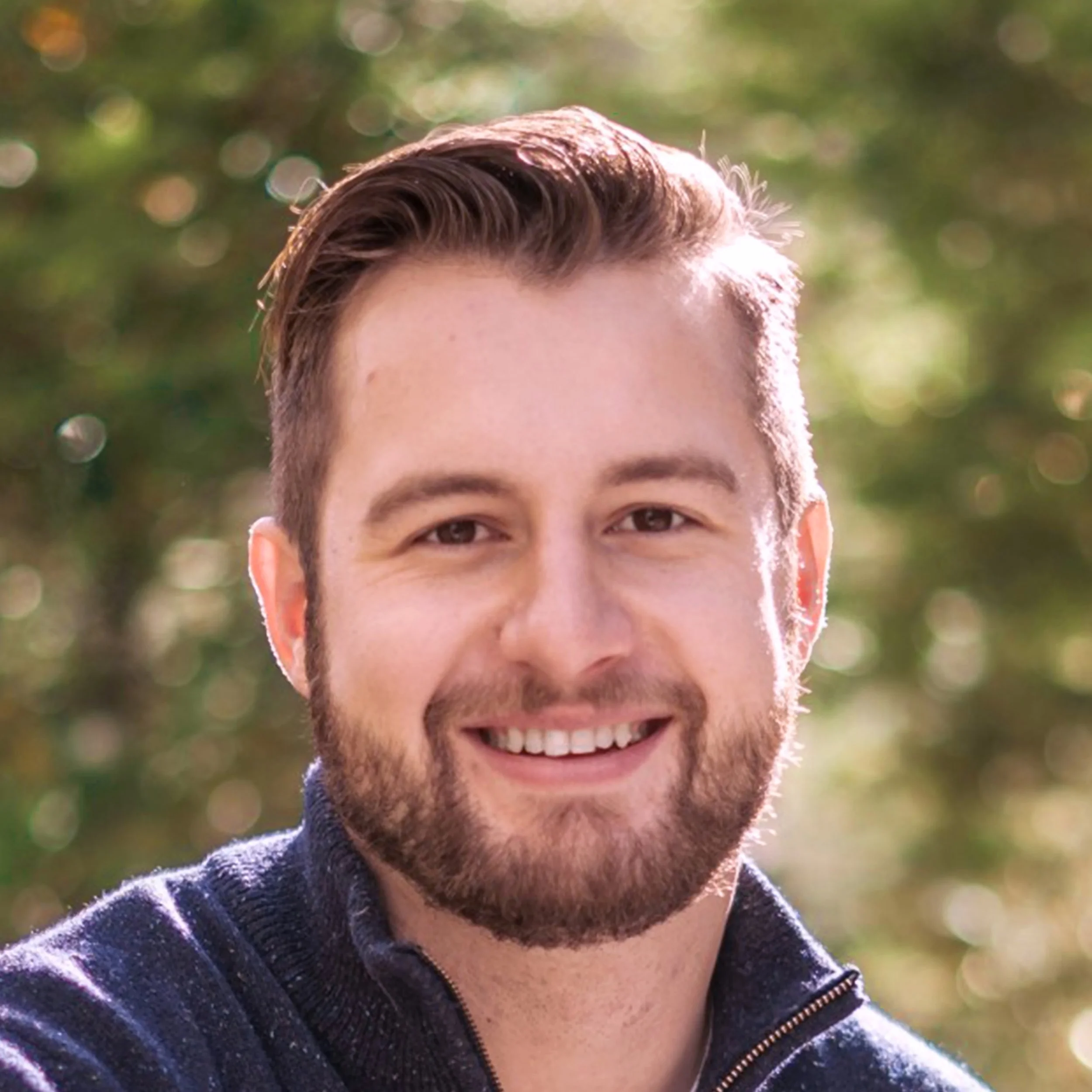 A smiling man with short, dark hair and a beard outdoors with blurred green trees in the background.