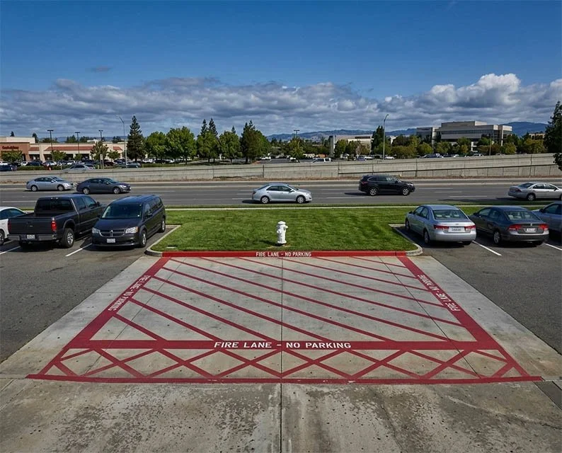 Empty parking lot with a designated fire lane area marked in red and white, fire hydrant nearby, with cars parked to the sides. In the background, a highway with moving vehicles and a commercial area with buildings and trees under a partly cloudy sky.