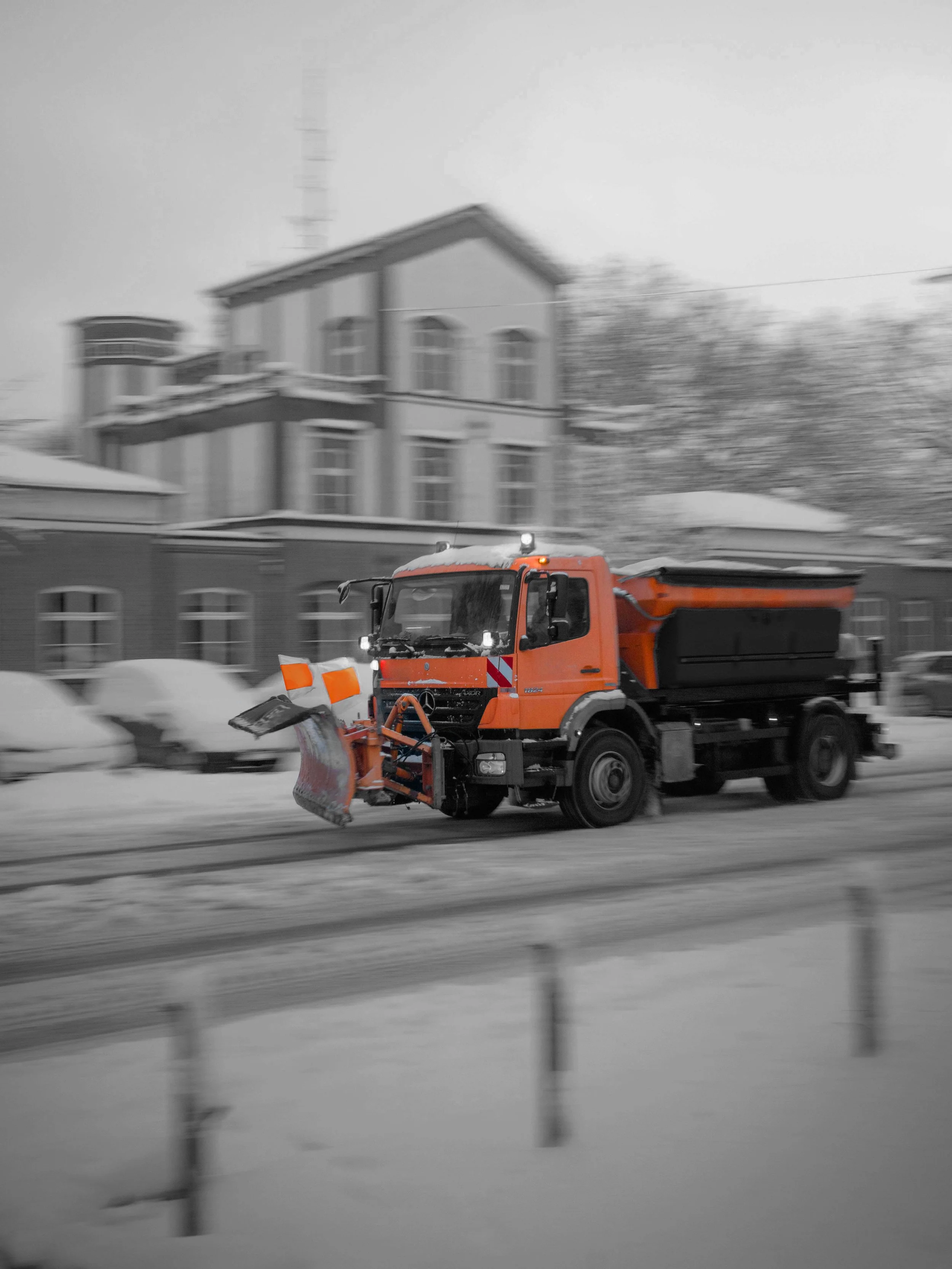 A snow plow truck clearing snow from the road in a winter scene, with a multi-story house and snow-covered ground in the background.