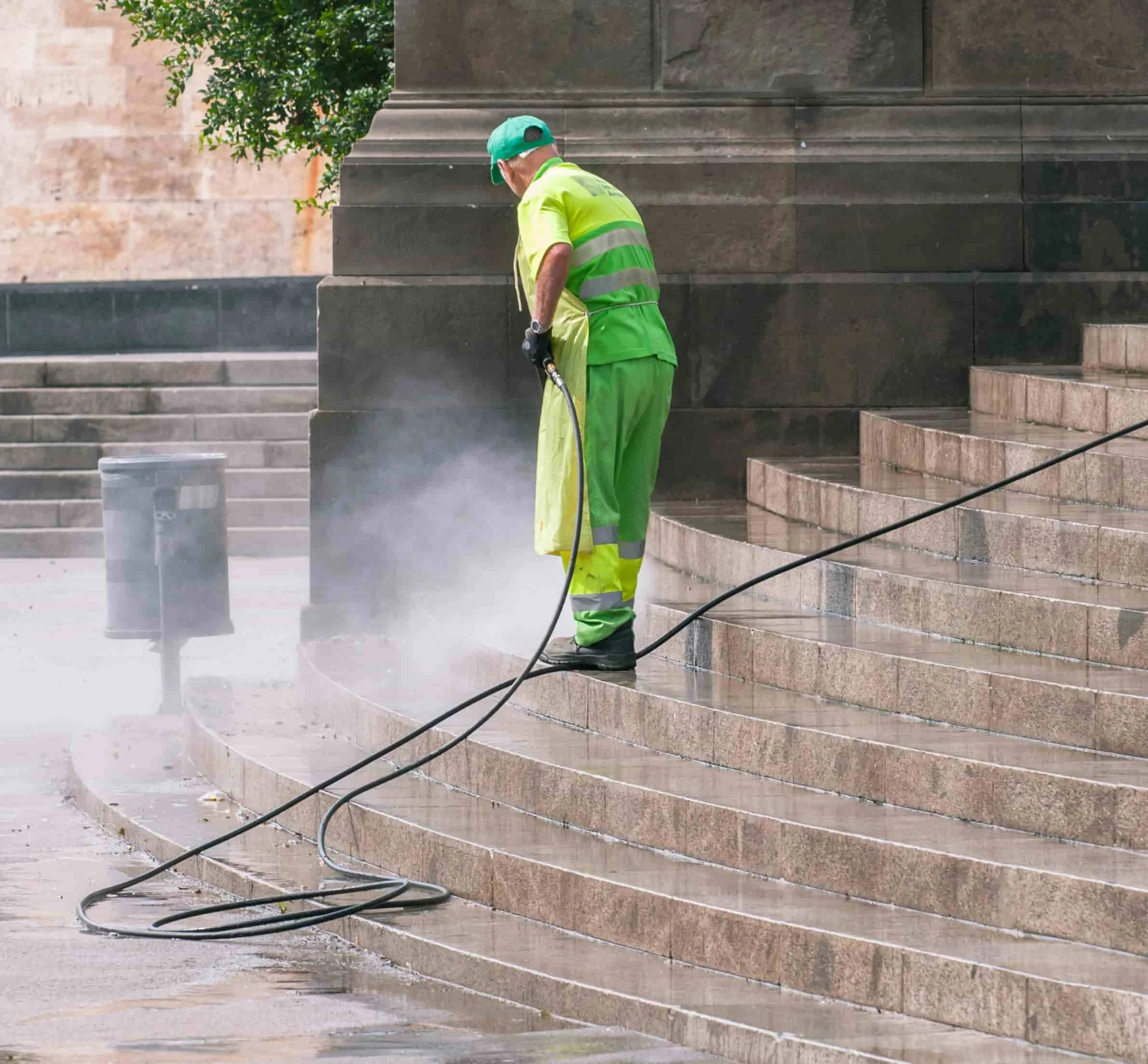 A worker wearing a green uniform, yellow vest, and green cap power washing stone steps in an outdoor area, with greenery visible in the background.
