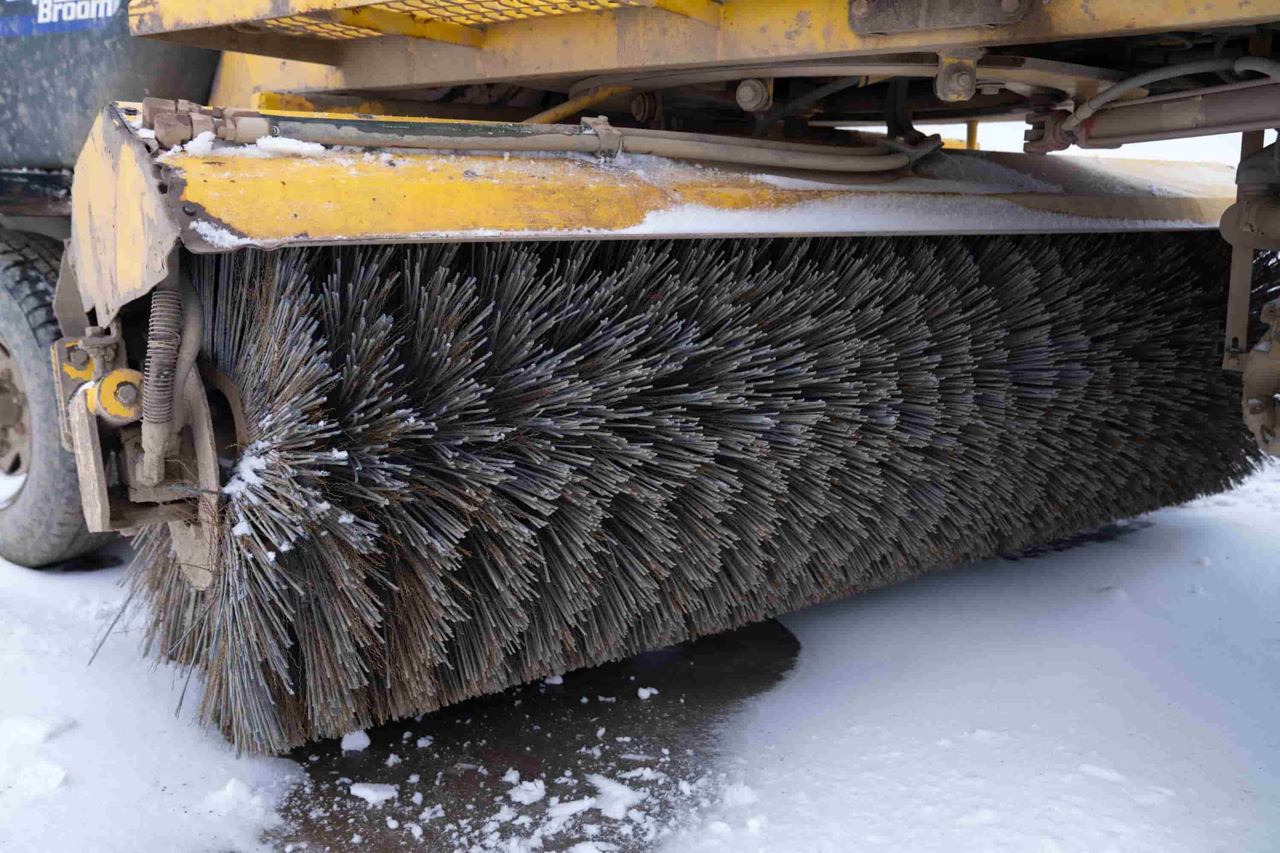 A large industrial snow plow with a rotating brush attachment clearing snow from a surface.