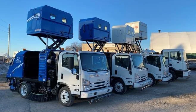 A line of street sweepers with large blue and gray boxes on top, parked outdoors on a gravel lot under a clear blue sky.