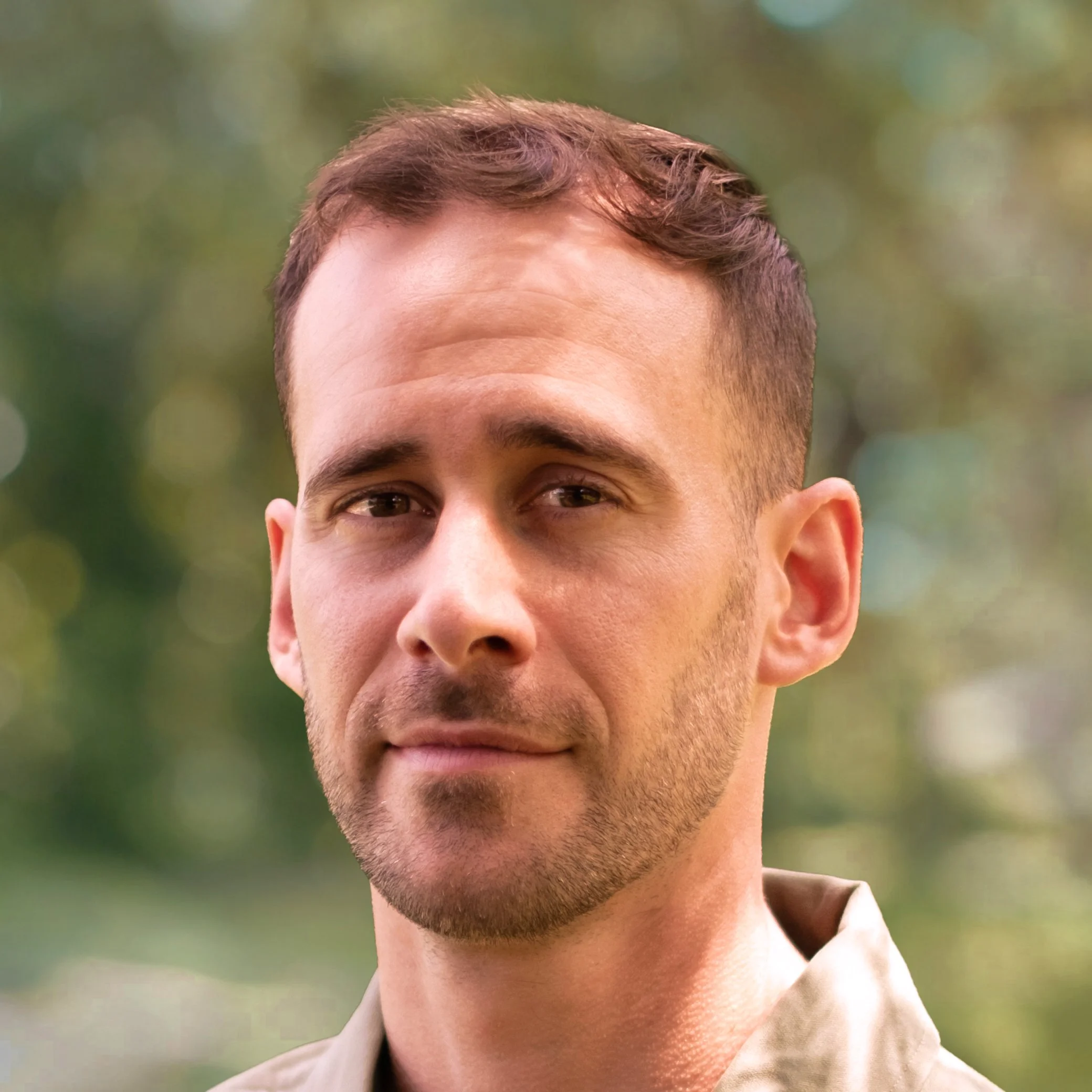 Close-up portrait of a young man with short brown hair and light facial hair, outdoors with blurred green background.