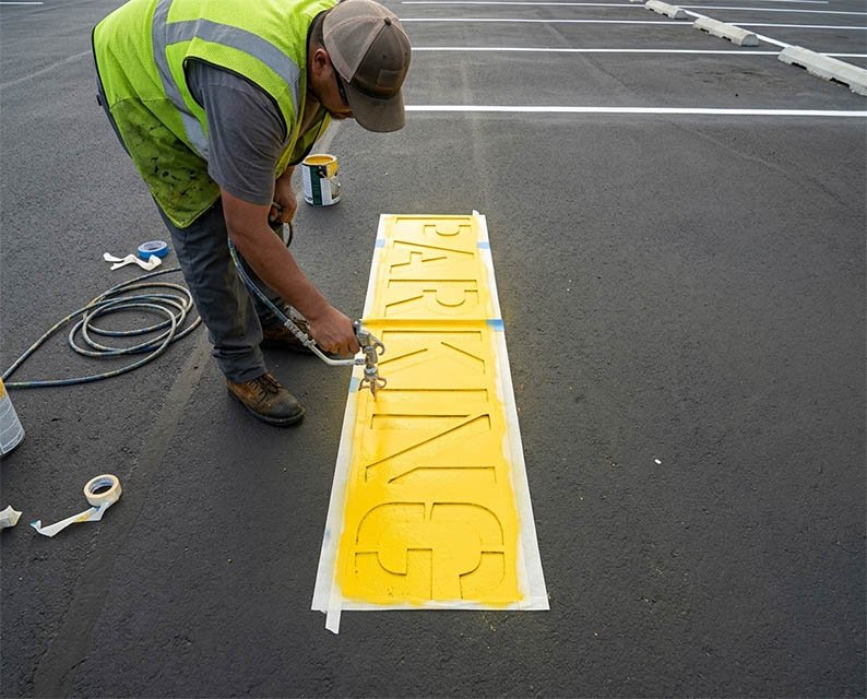A worker in a yellow safety vest and cap spray painting yellow letters onto a parking lot pavement, creating a new sign.