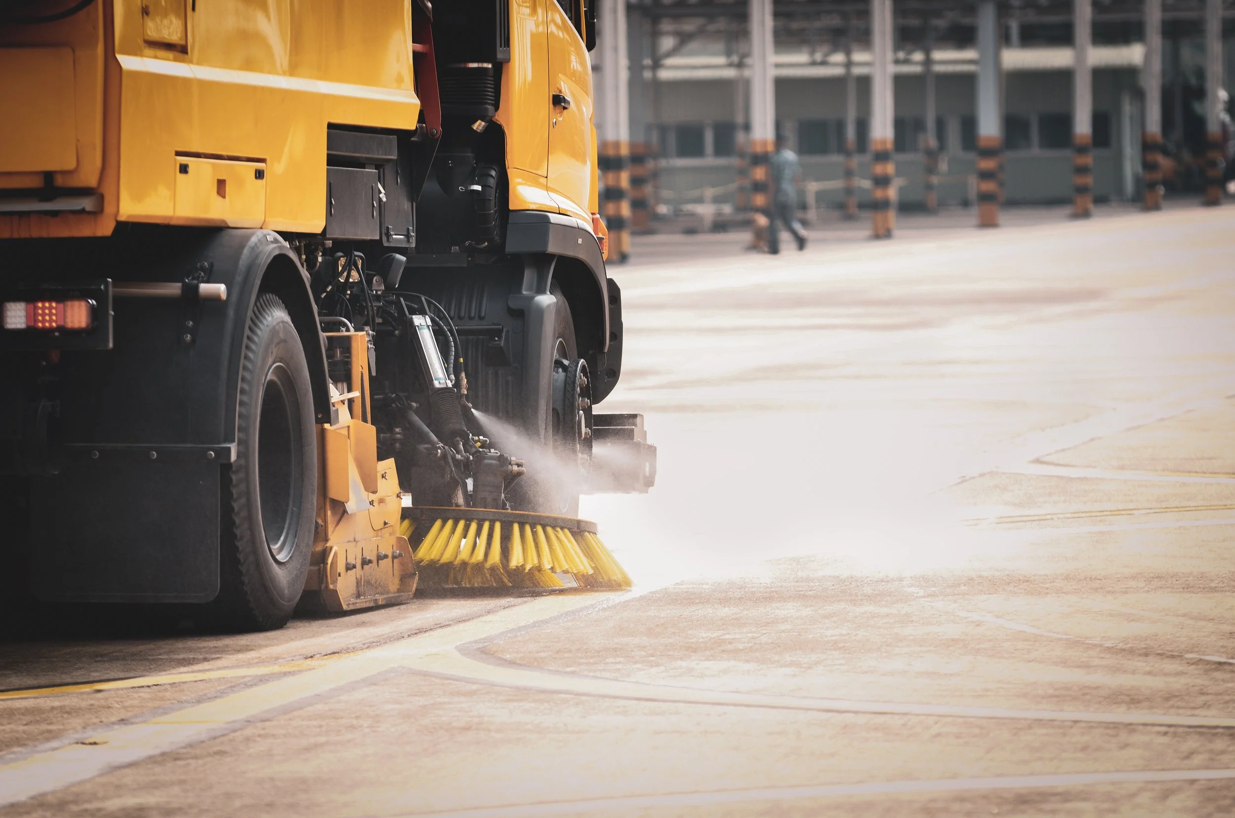 A street cleaning machine with a yellow body and a large rotating brush cleaning the pavement in an urban area.