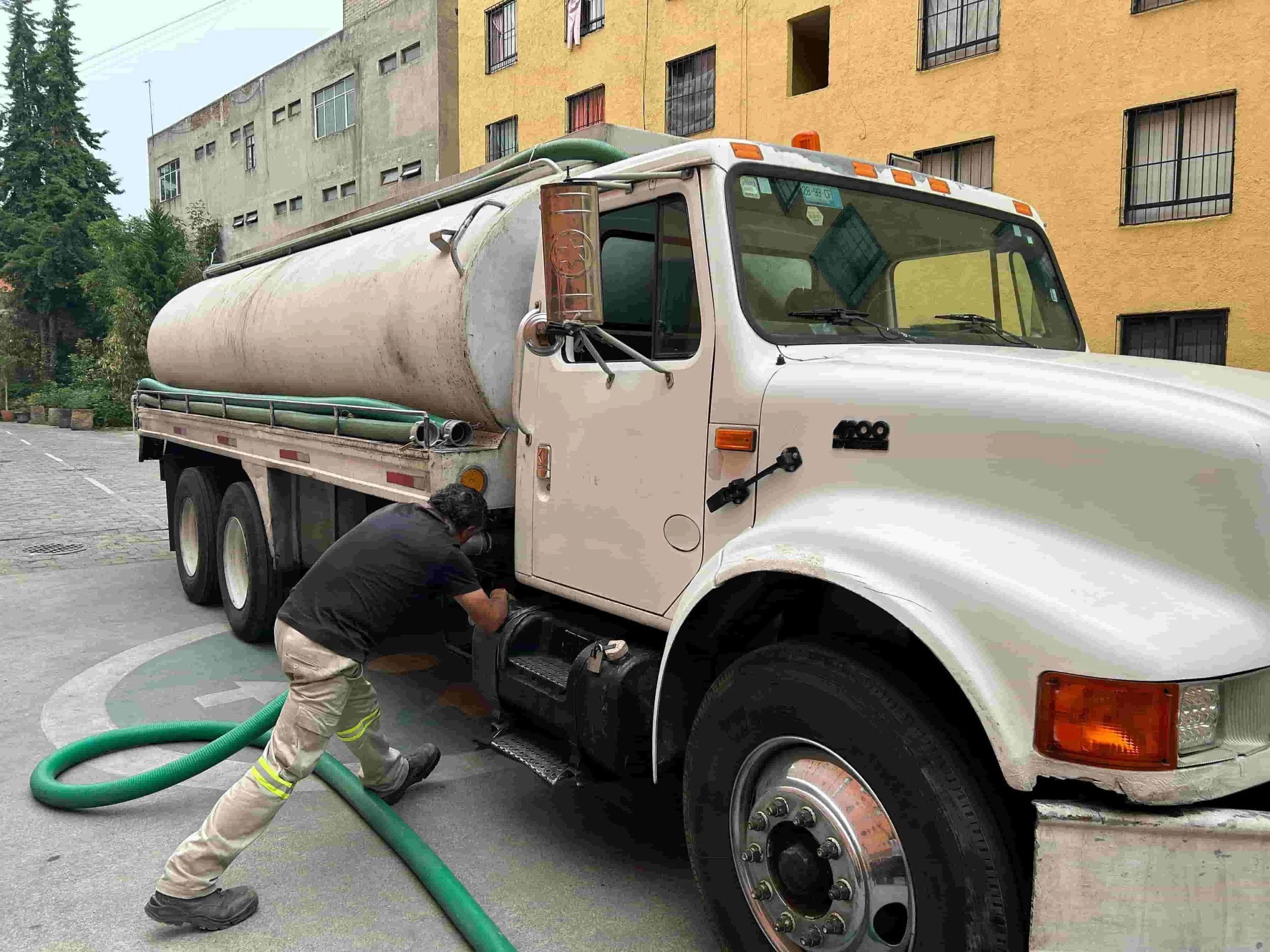 A worker in yellow reflective pants is adjusting a green hose connected to a white tanker truck parked in a residential area with multicolored buildings.