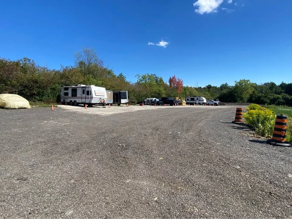 A parking lot with several RVs and trucks, orange and black striped traffic cones, and a small patch of greenery with yellow flowers under a blue sky with a few clouds.