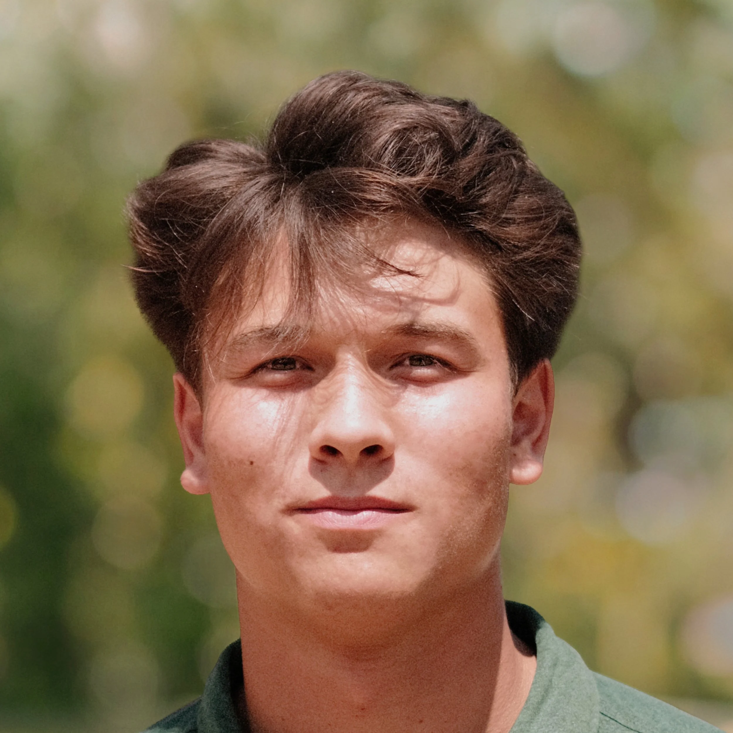 A young man with brown hair outdoors, wearing a green shirt, with a blurred green and yellow background.