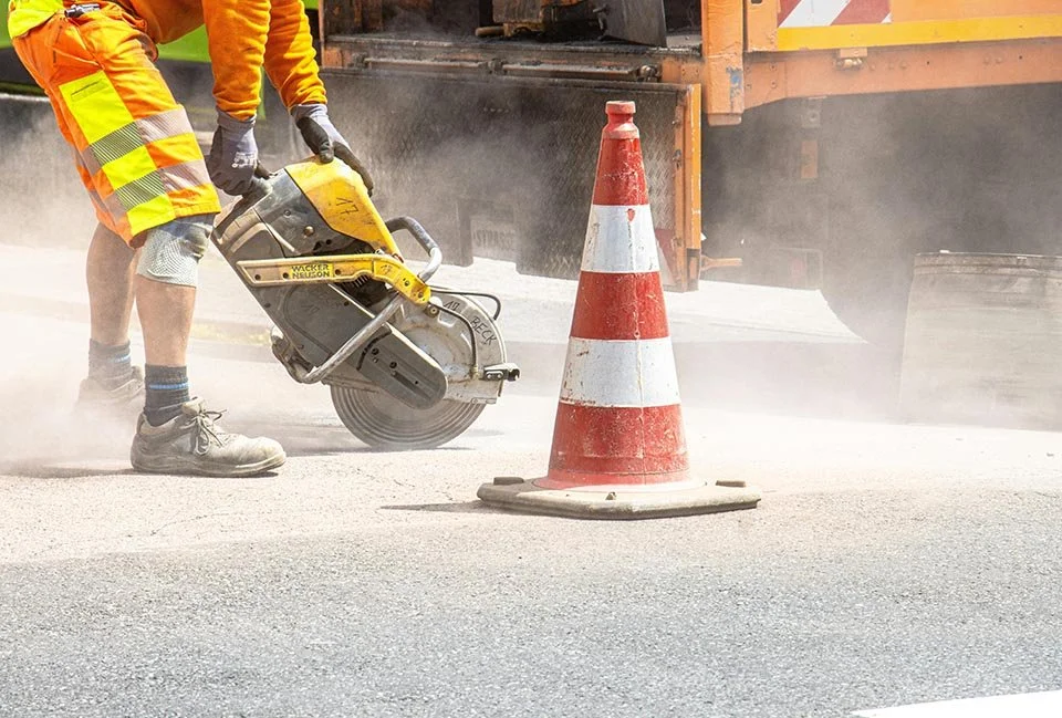 Worker cutting pavement on road near orange traffic cone and construction vehicle.
