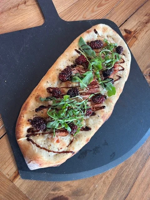 Flatbread pizza topped with blackberries, greens, and drizzled with balsamic glaze on a black serving board.