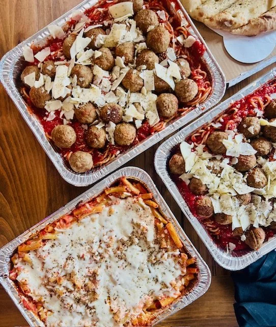 Three aluminum trays of lasagna with meatballs, cheese, and tomato sauce on a wooden table.