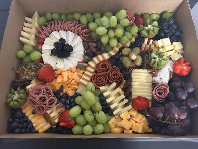 A box of assorted sliced cheeses, rolled meats, green and purple grapes, strawberries, blueberries, and other fruits arranged for a cheese platter