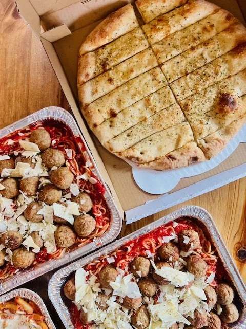 A cheese pizza in a box and two trays of spaghetti with meatballs topped with cheese and marinara sauce on a wooden table.