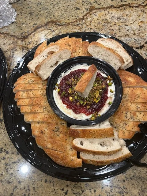 A round black platter with sliced baguette bread surrounding a small bowl of creamy spread topped with raspberry jam, chopped pistachios, and a small piece of bread.